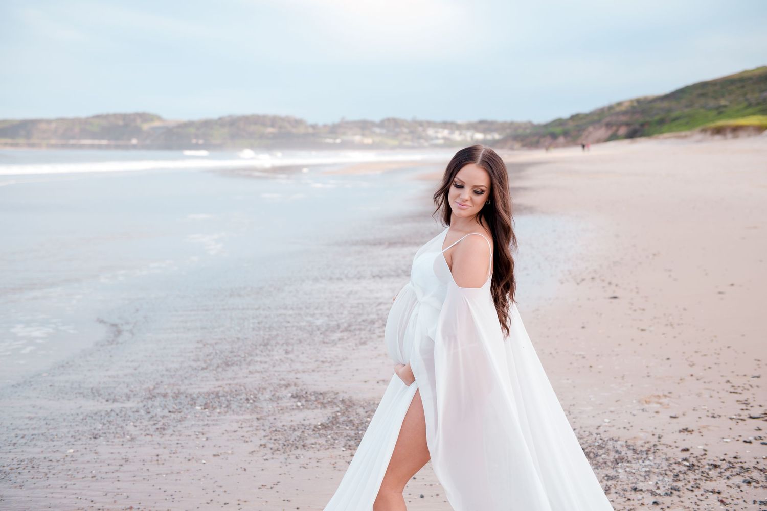 A pregnant person in a flowing white dress walks along a scenic beach shore with mountains in the background.