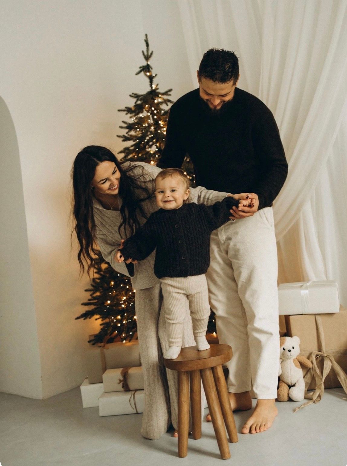 Family with baby standing on stool near decorated Christmas tree with gifts and teddy bear in white room.
