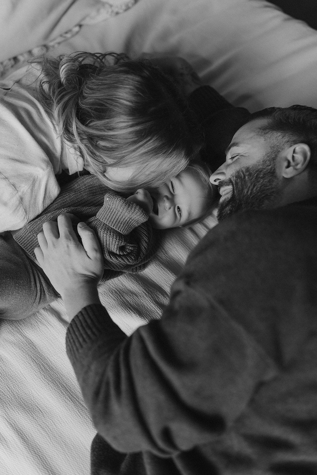 Black and white photo of parents lying in bed smiling with their baby between them.