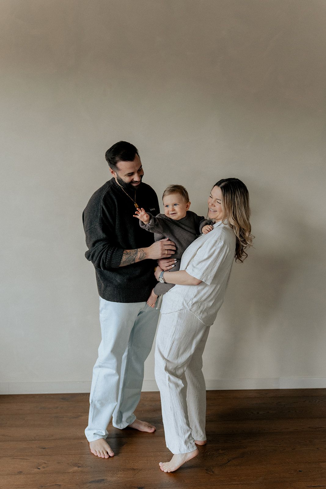 Smiling family with tattooed father holding toddler boy while pregnant mother looks on.