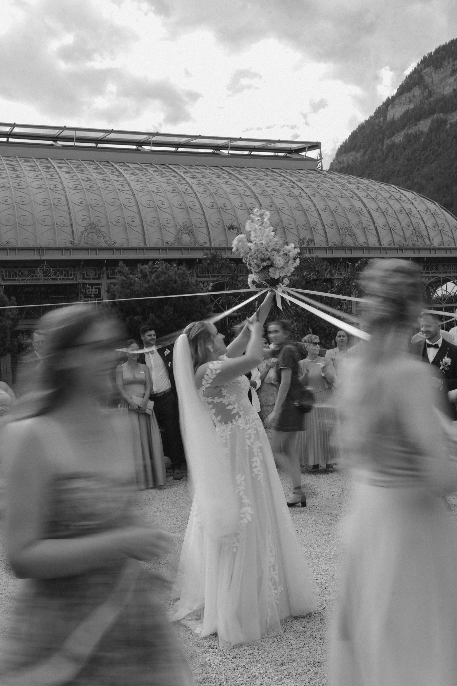 Bride tosses bouquet surrounded by blurred moving guests outside a glass pavilion in black and white.