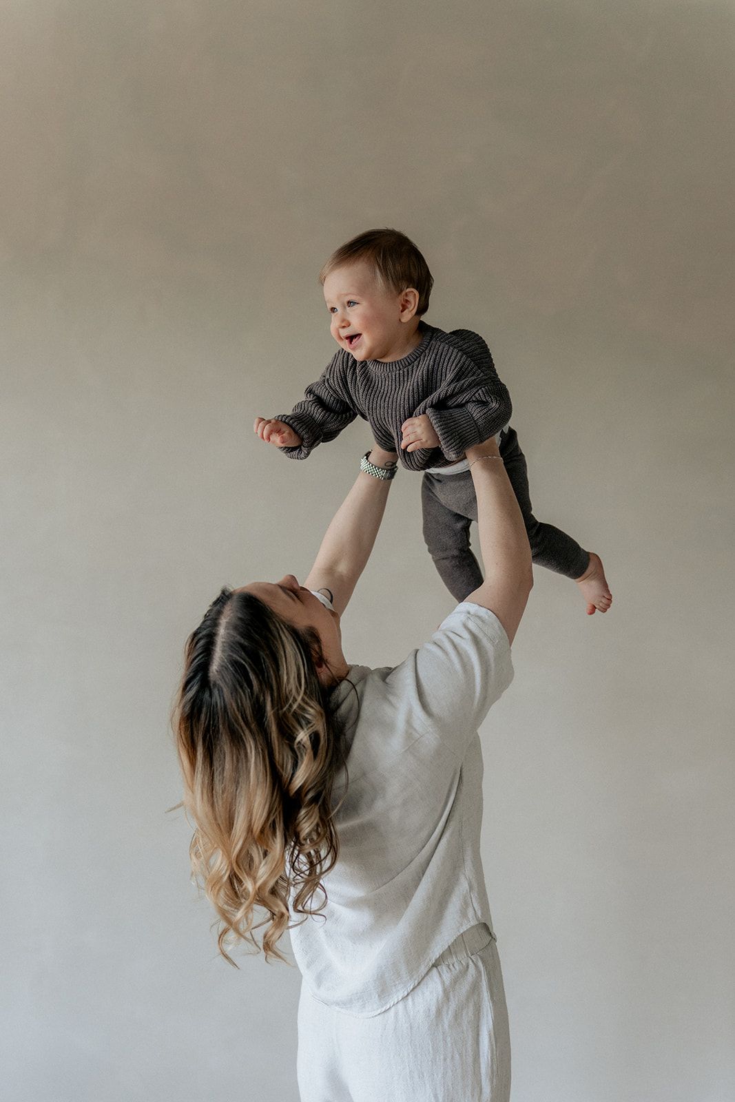Mother in white outfit lifting happy baby overhead against neutral beige wall in bright room.