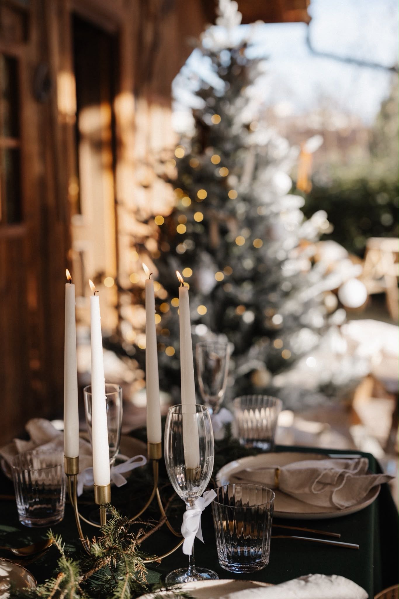 Elegant winter wedding table setting with white candles, crystal glasses, and a snow-dusted Christmas tree in the background.