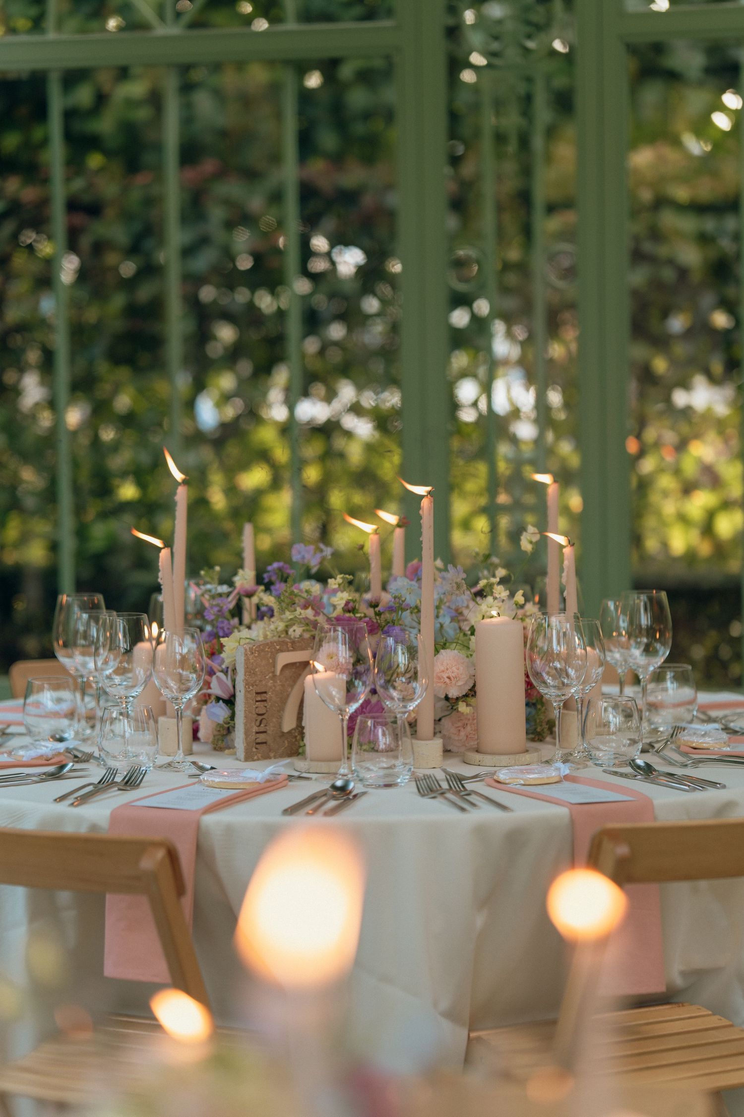 Romantic wedding table setting with tall taper candles, pink florals, and crystal glassware in greenhouse venue.