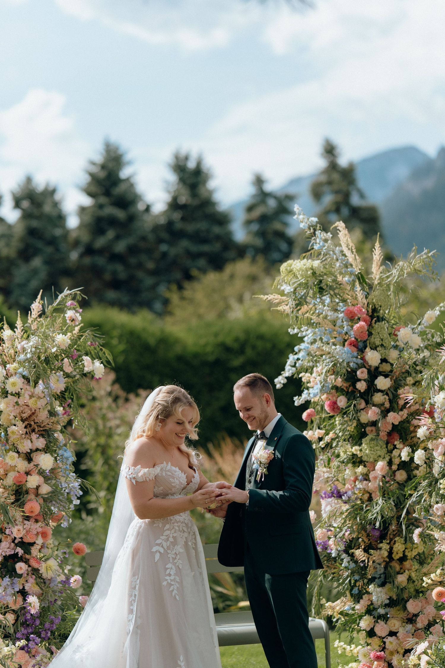 Couple exchanges rings at a floral arch ceremony with colorful blooms and mountain views behind.