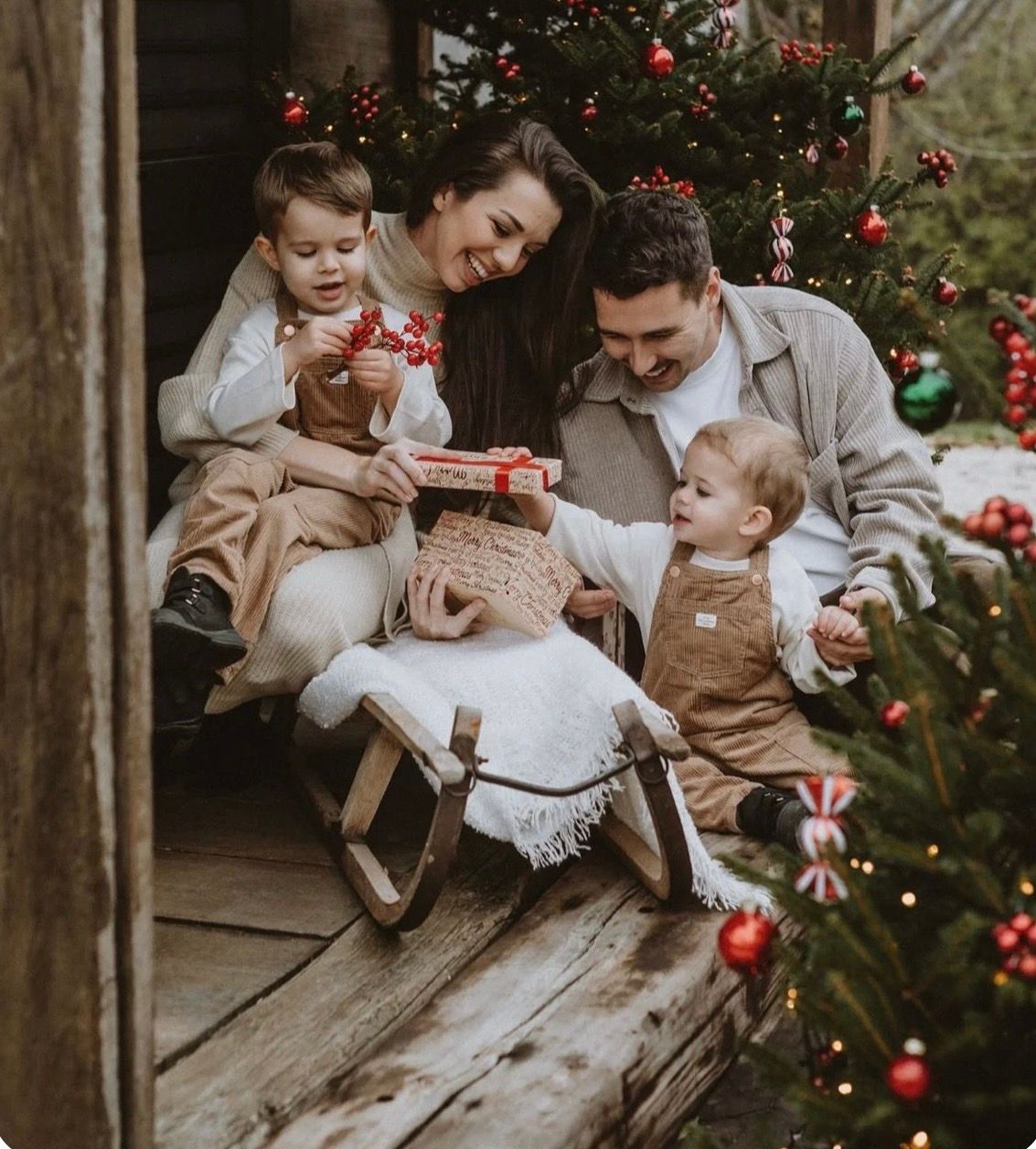 Family of four celebrating Christmas on a rustic porch with festive decorations and a sled.