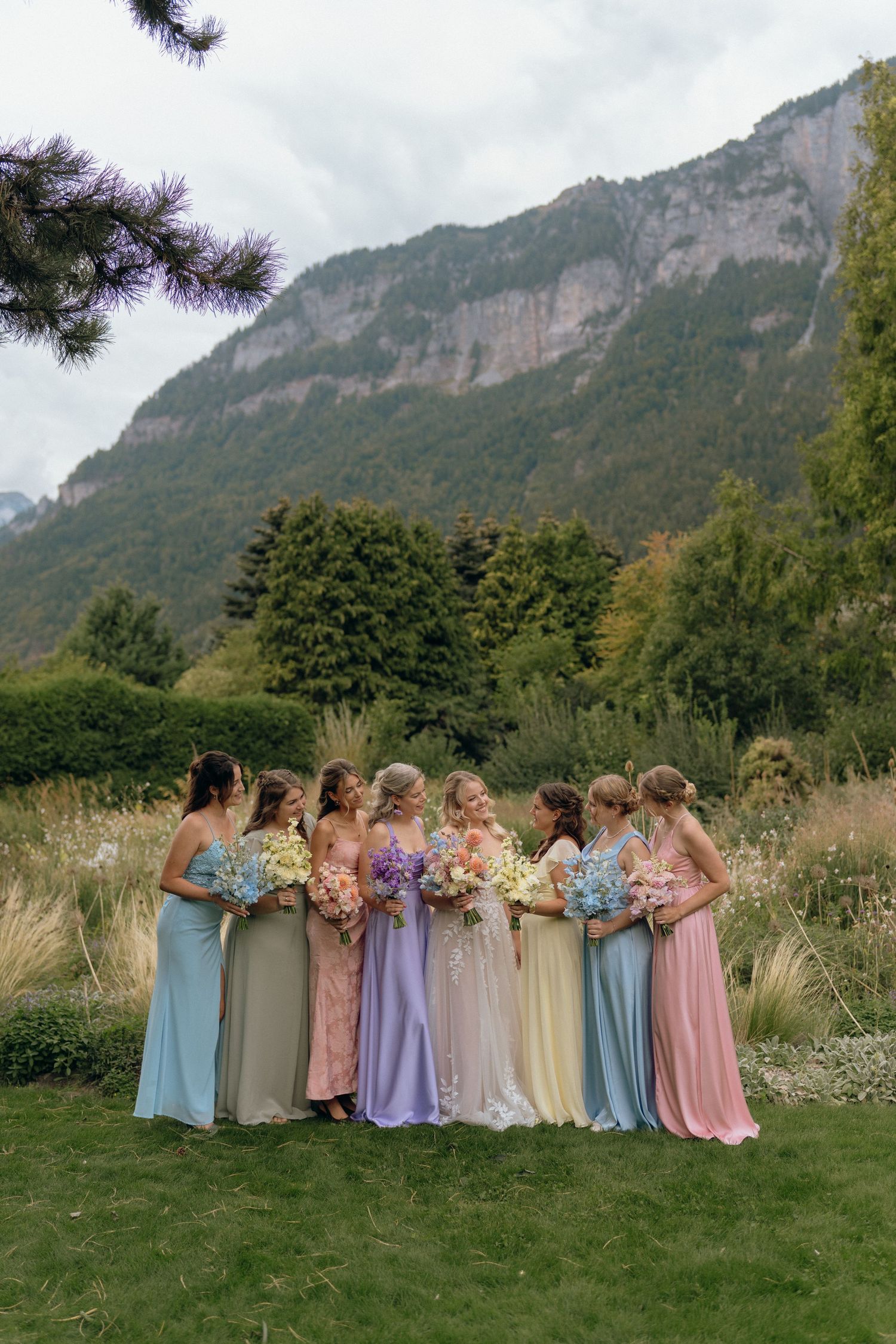 Bride stands with seven bridesmaids in mismatched pastel gowns holding bouquets before mountains.