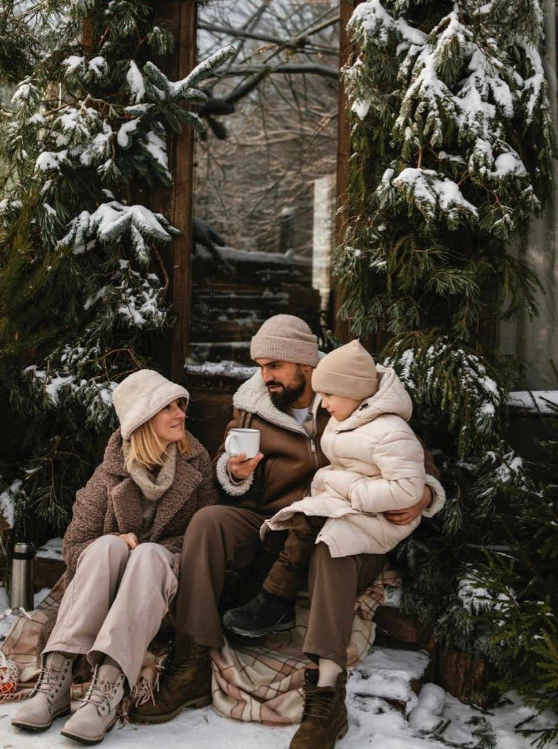 Family in winter hats and coats sitting outdoors in snow, sharing warm drinks beside snow-covered evergreen trees.