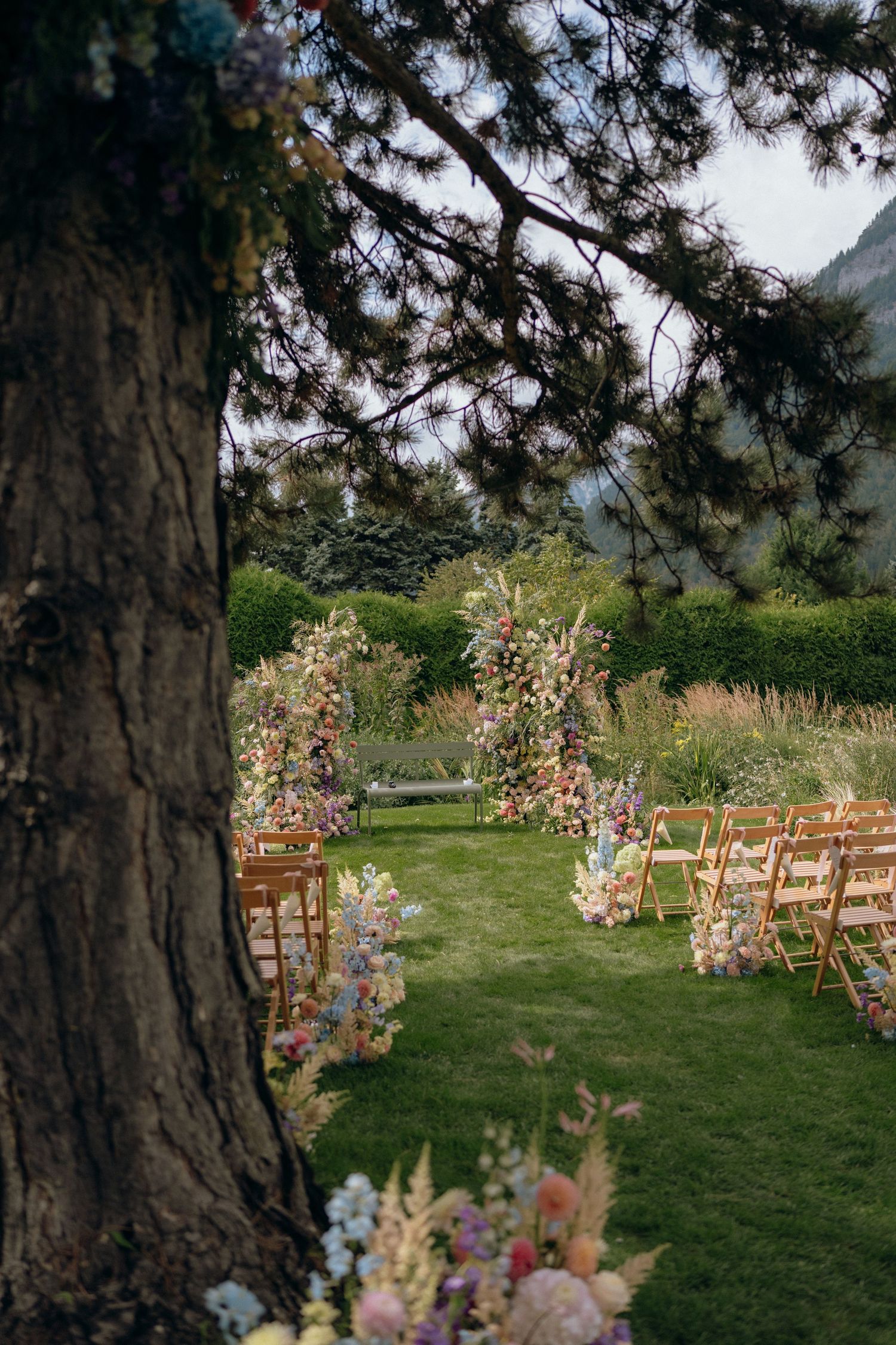 Elegant outdoor wedding ceremony setup with colorful floral arches on a green lawn near mountains.