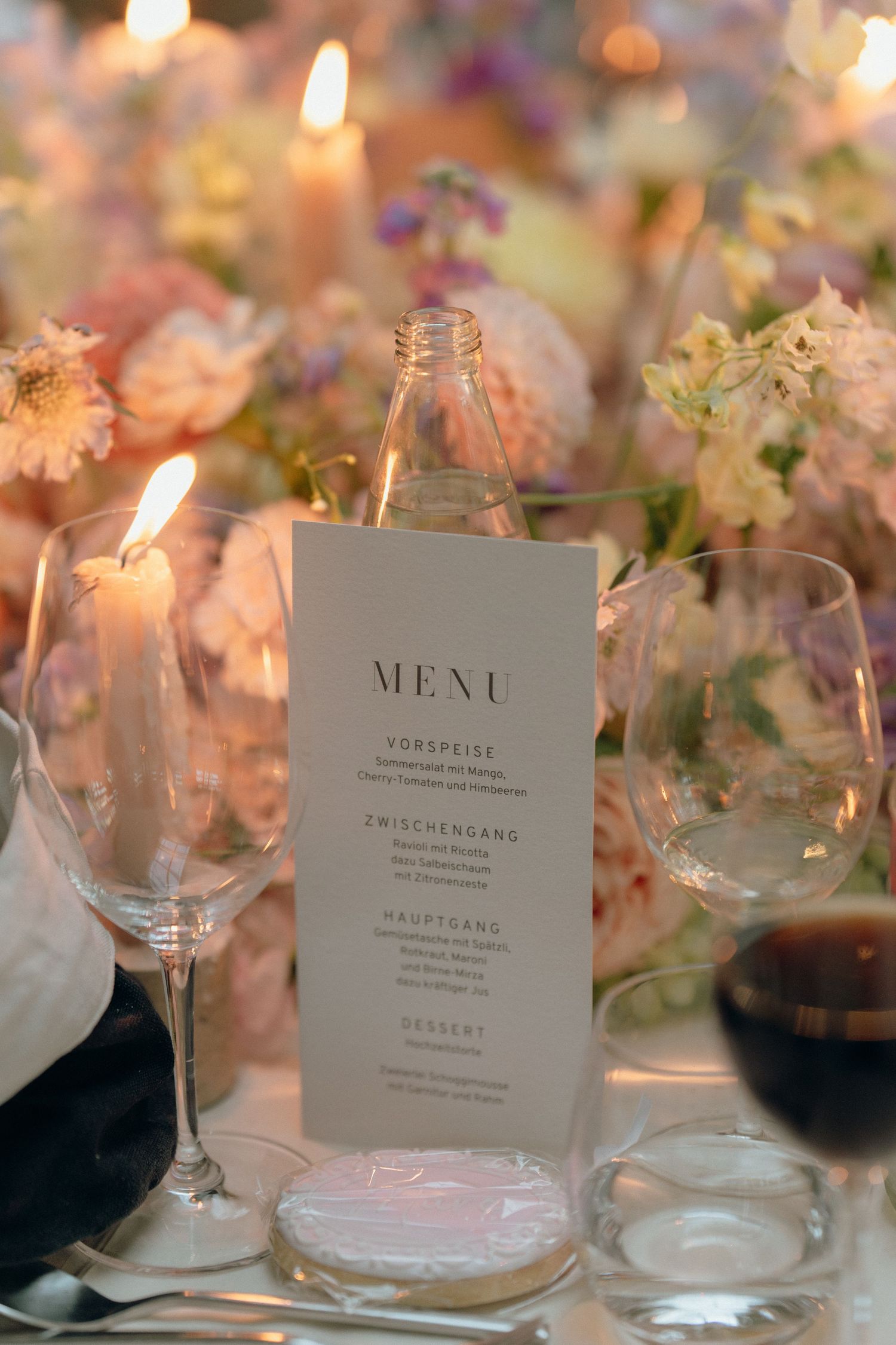 Elegant wedding menu card surrounded by soft pink florals, candles, and wine glasses on reception table.