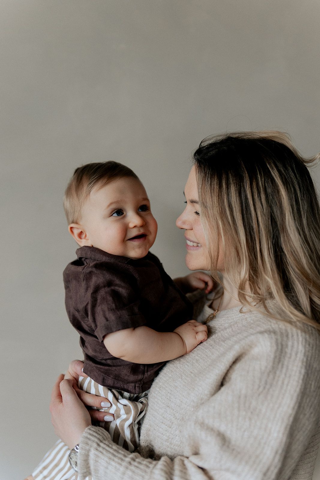 Joyful baby in dark outfit smiling at blonde mother during an indoor family portrait session.