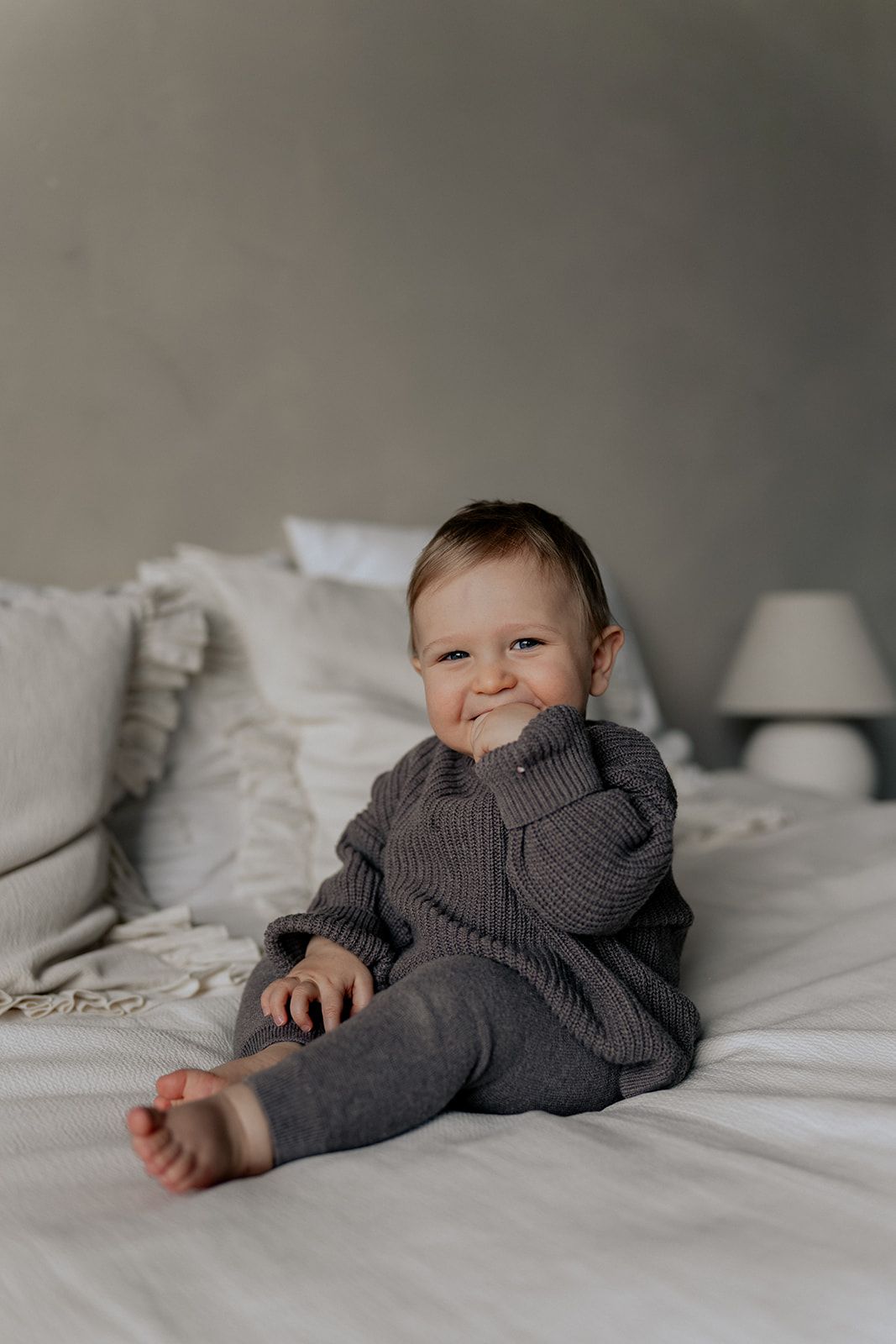 Adorable baby in grey knit outfit sitting on white bed grinning.