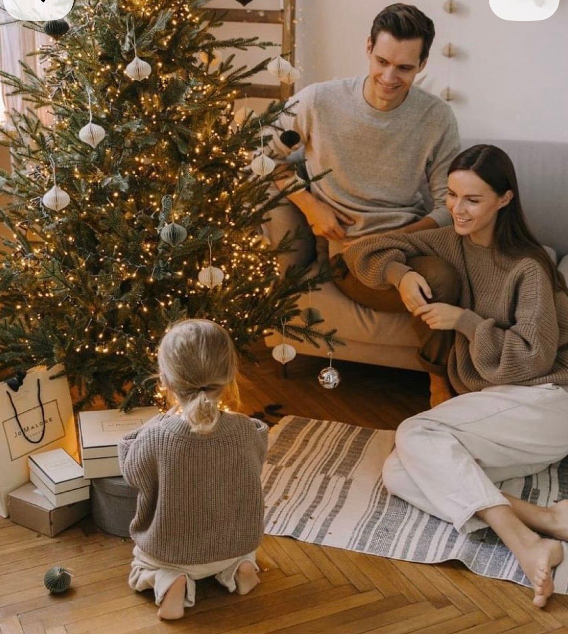 Family decorating Christmas tree together, parents and child sitting on floor with ornaments in cozy living room.