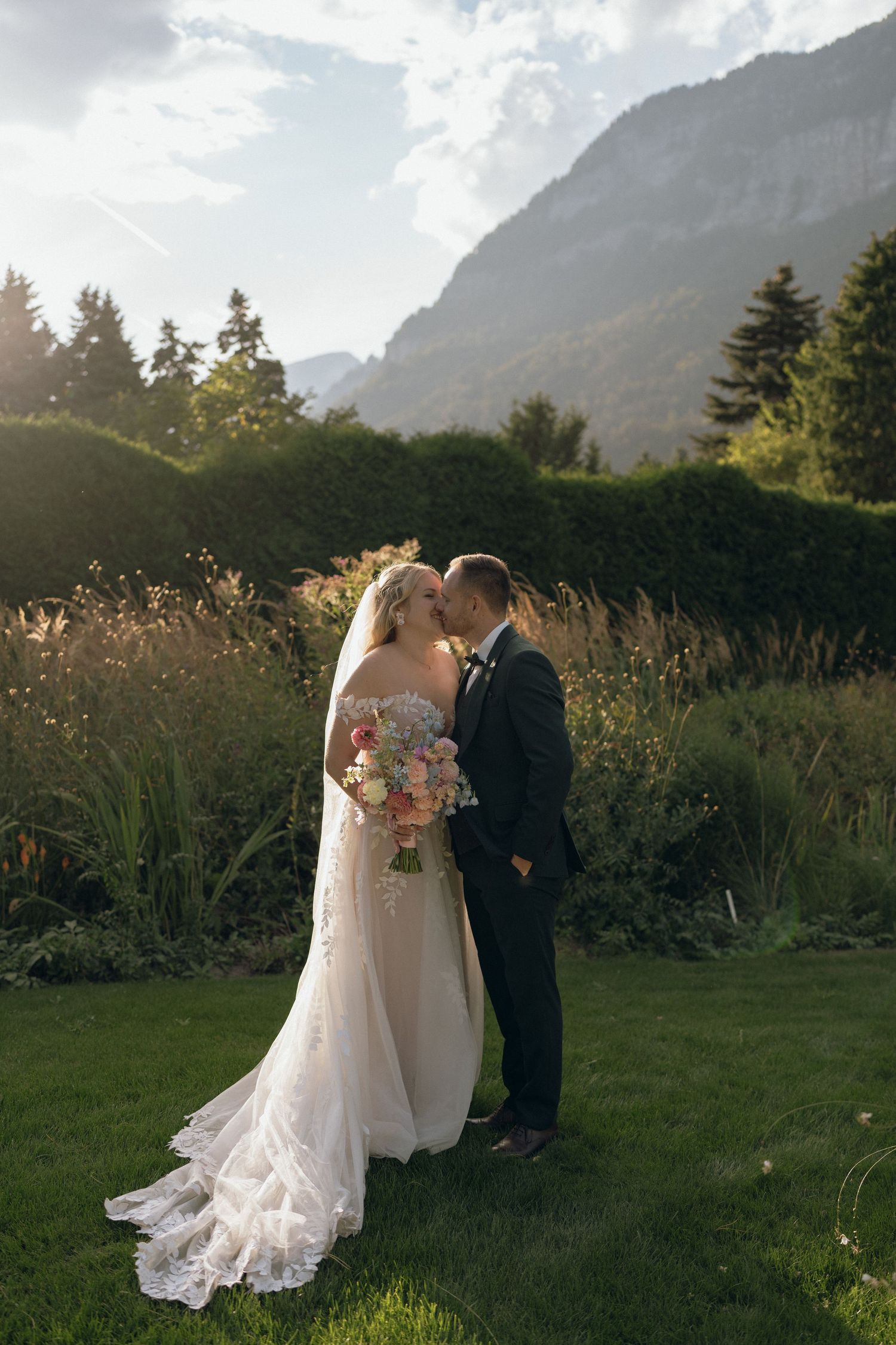 Bride and groom share a romantic kiss in lush garden with dramatic mountain backdrop at sunset.