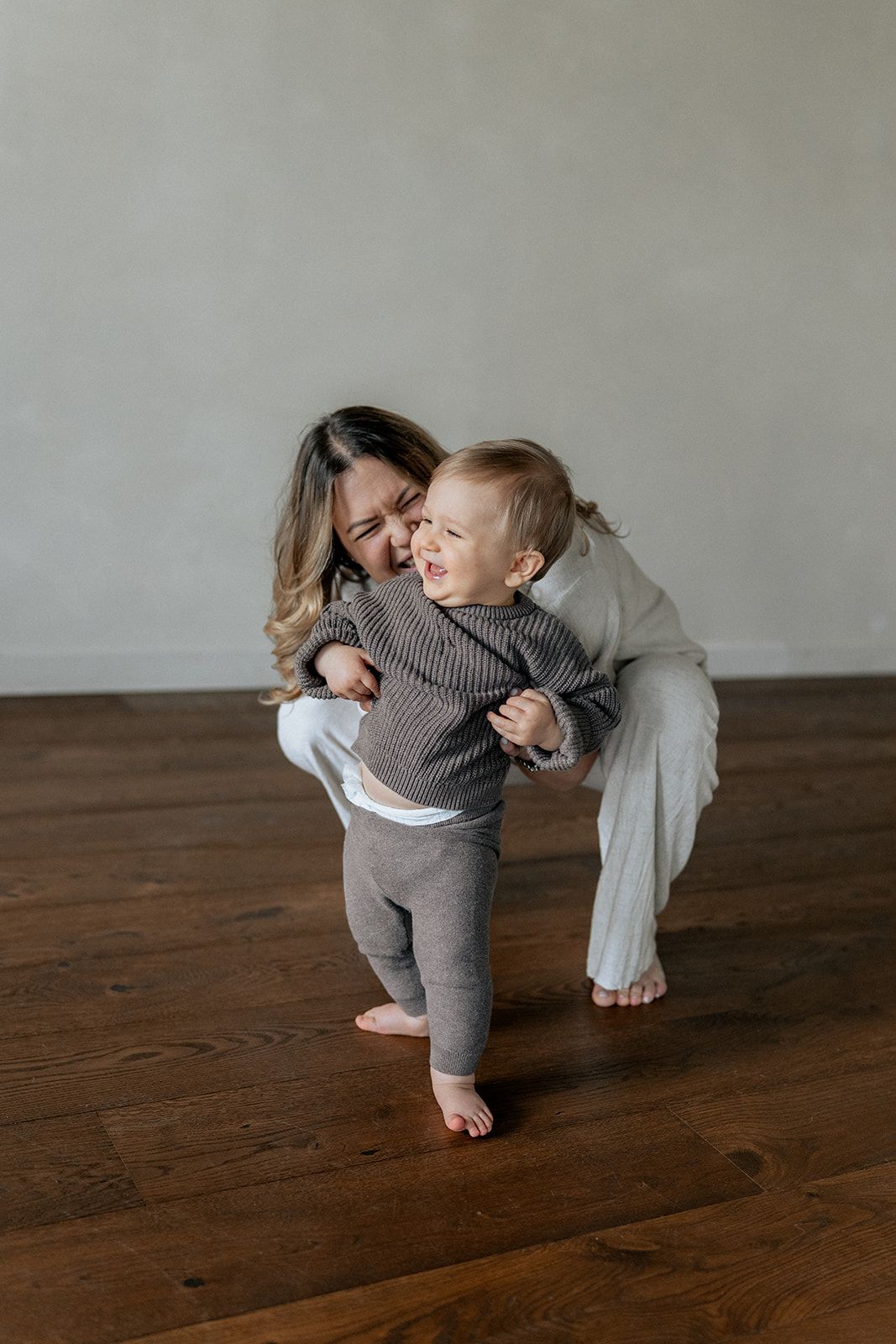 Mother in gray outfit hugging laughing barefoot toddler in knit romper on dark hardwood floor.