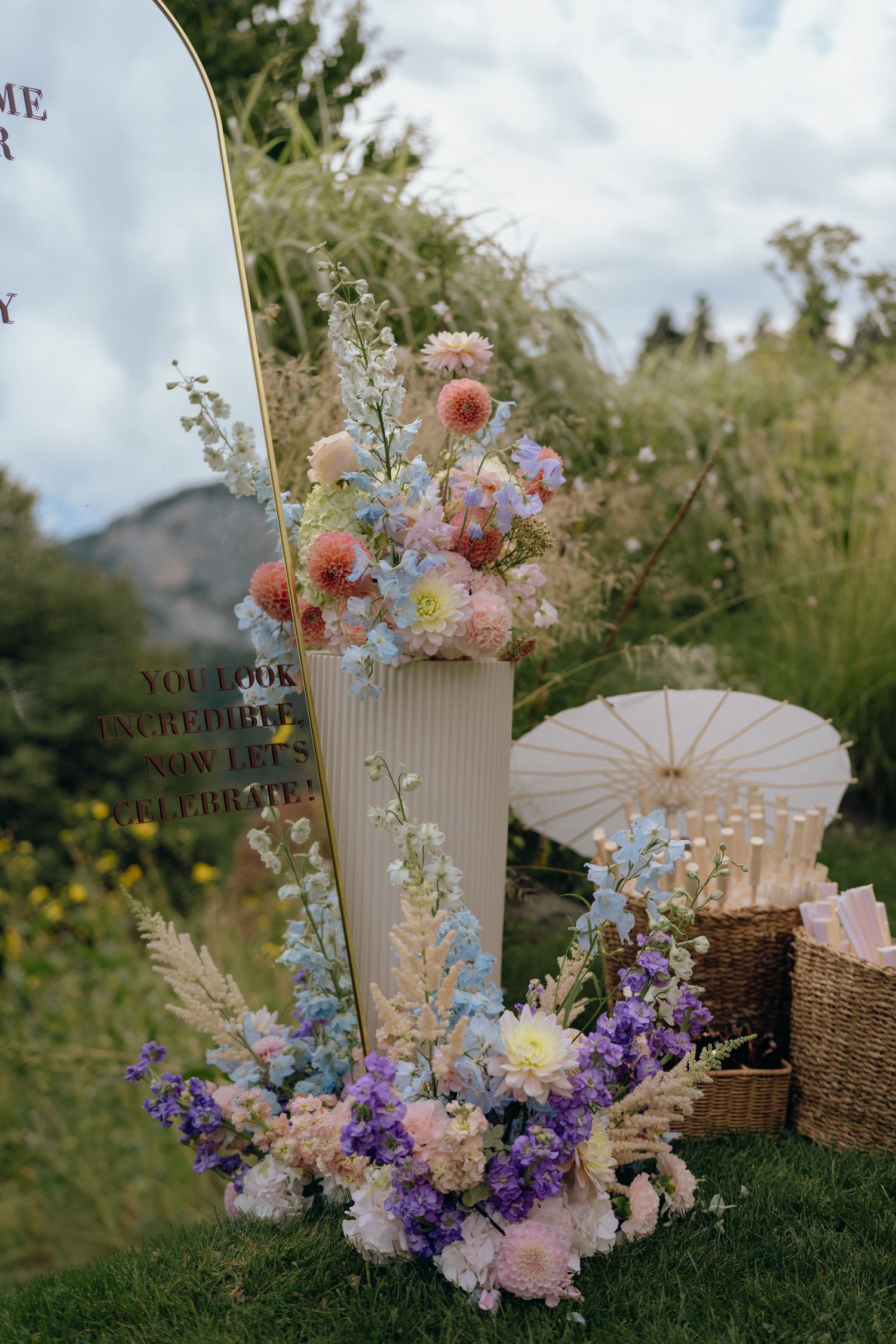 Pastel wedding welcome display with colorful dahlia and wildflower arrangements, parasols, and gold mirror sign.