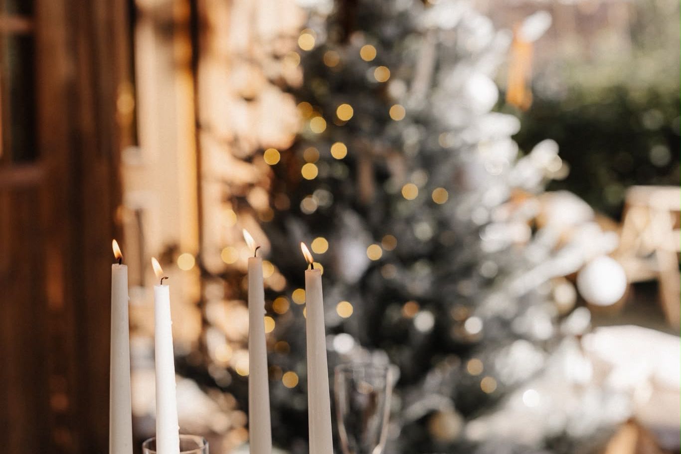 White candles glowing in front of a snow-flocked Christmas tree with golden bokeh lights in a festive holiday setting.
