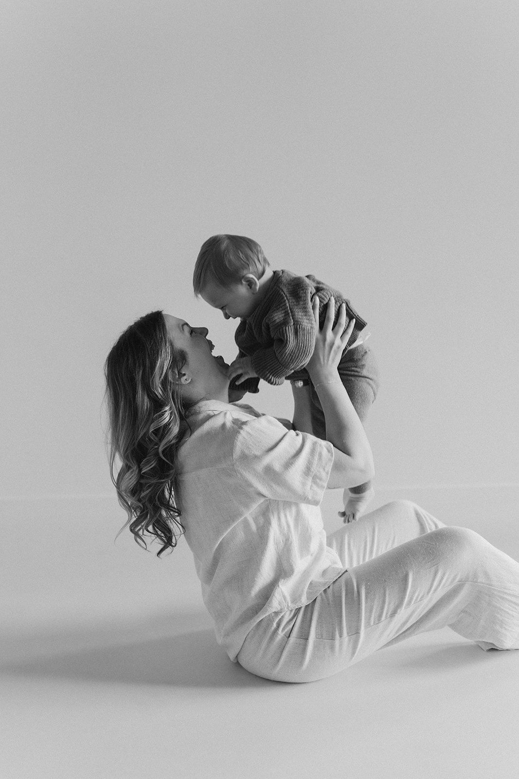 Black and white photo of mother sitting on floor holding baby up to her face.