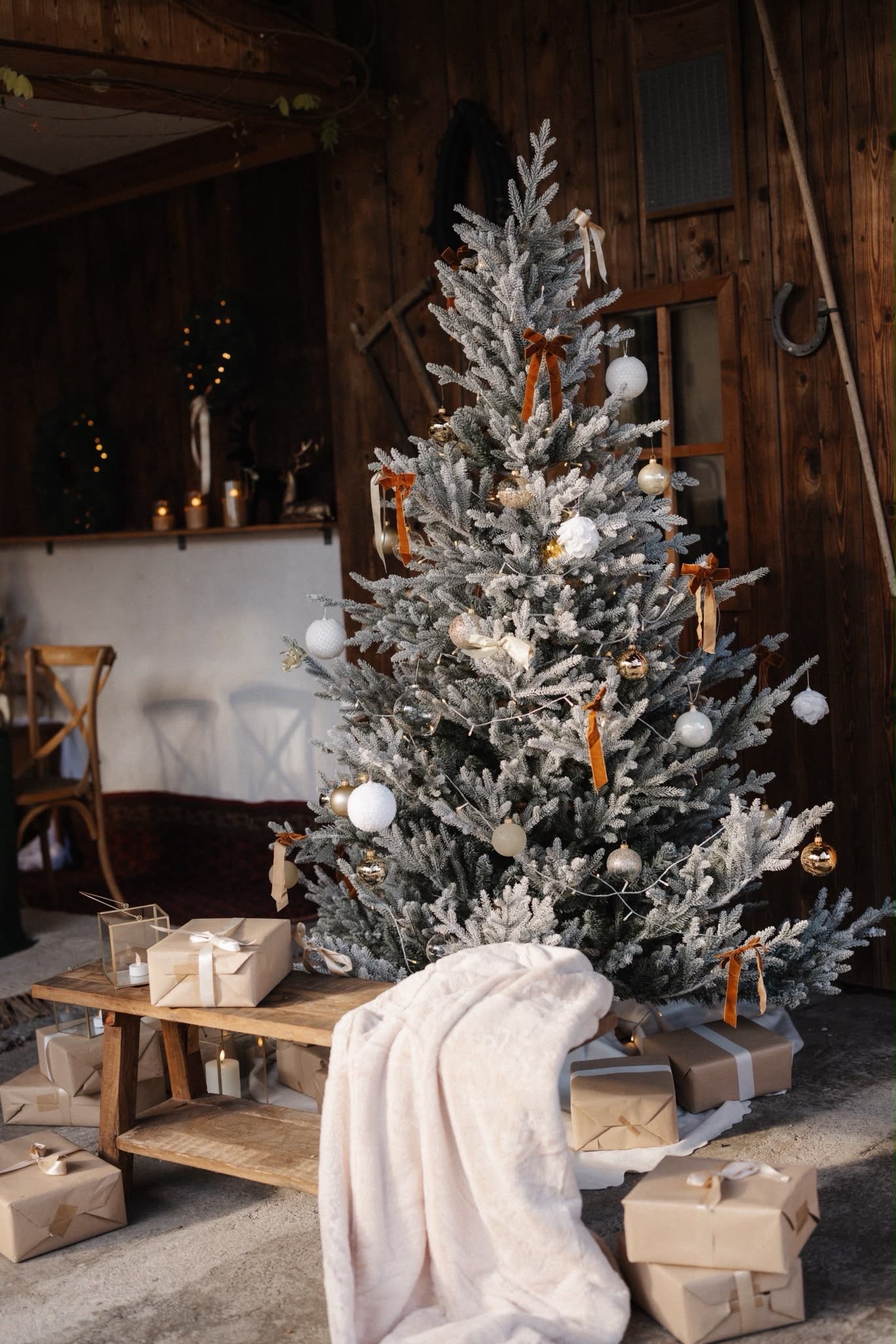 Snowy Christmas tree with white ornaments and gifts on wooden bench in rustic interior setting.