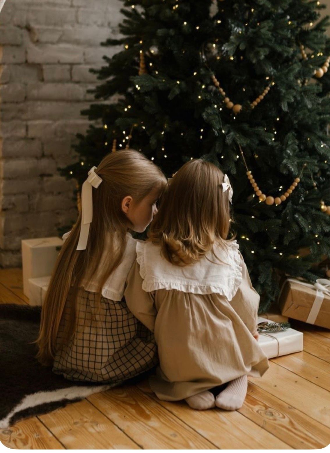 Two young girls in vintage dresses sit together admiring a decorated Christmas tree surrounded by gifts.