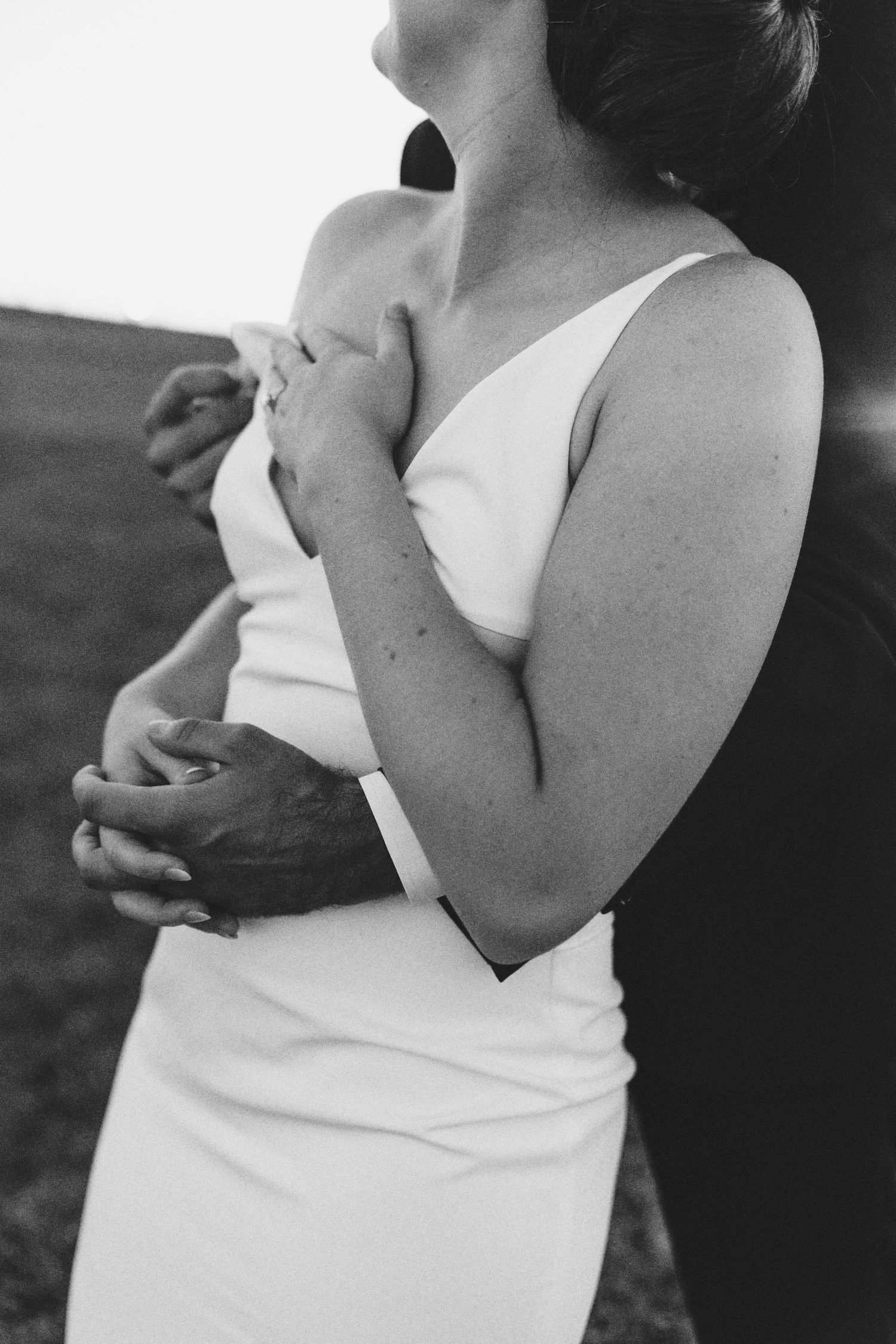 Black and white photo of a couple embracing, woman in white one-shoulder dress with man's arms wrapped around her.