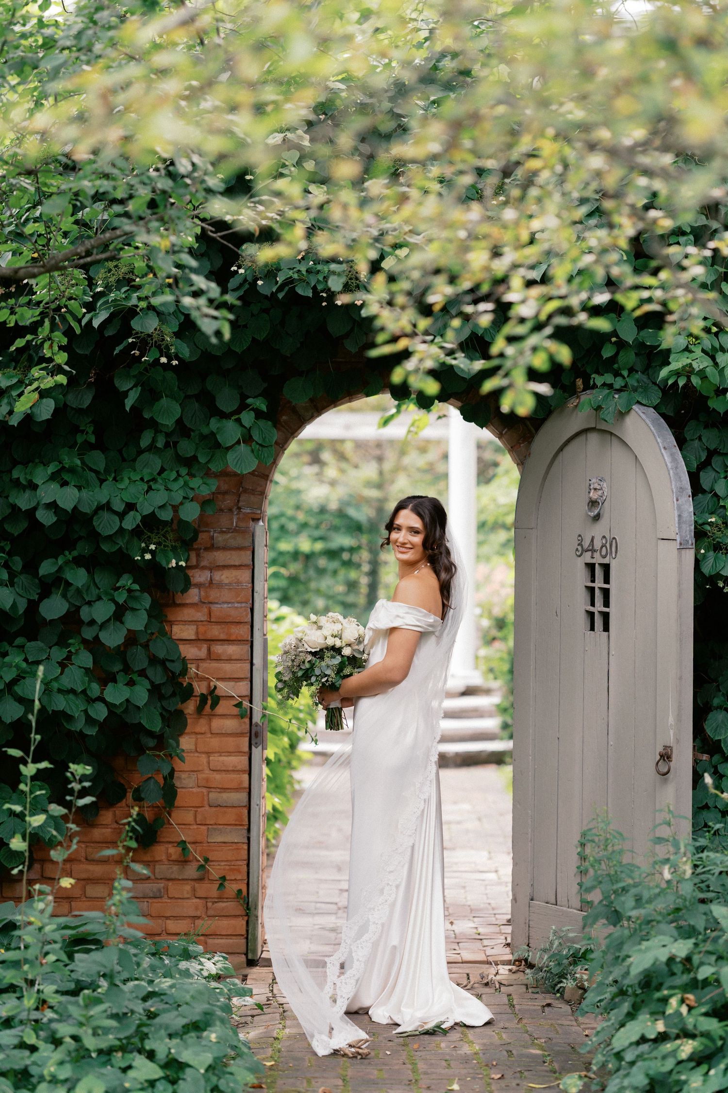 A bride in an off-shoulder white gown stands in a garden archway surrounded by lush greenery and brick walls.