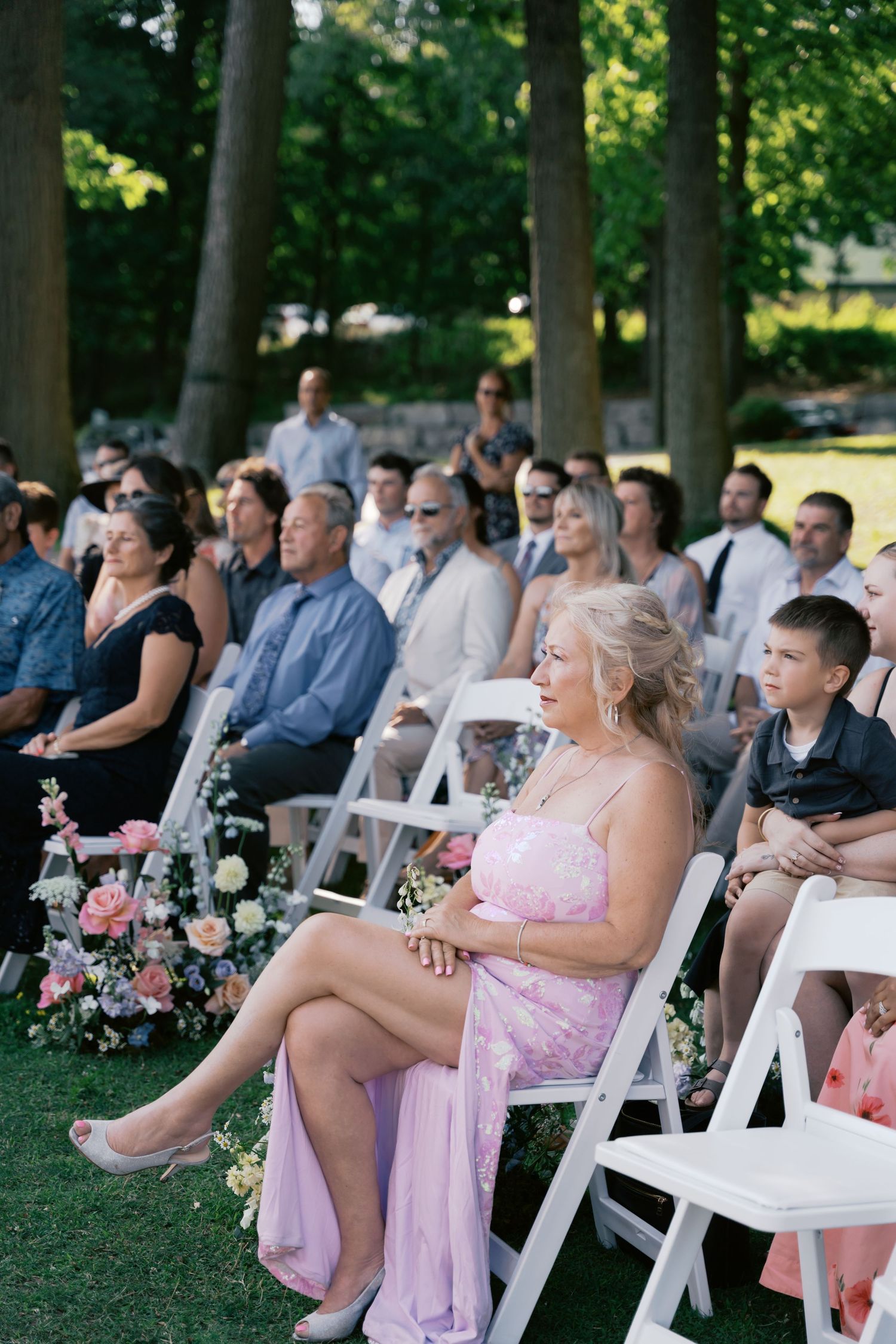 Wedding guests seated in white chairs under shaded trees during outdoor ceremony.