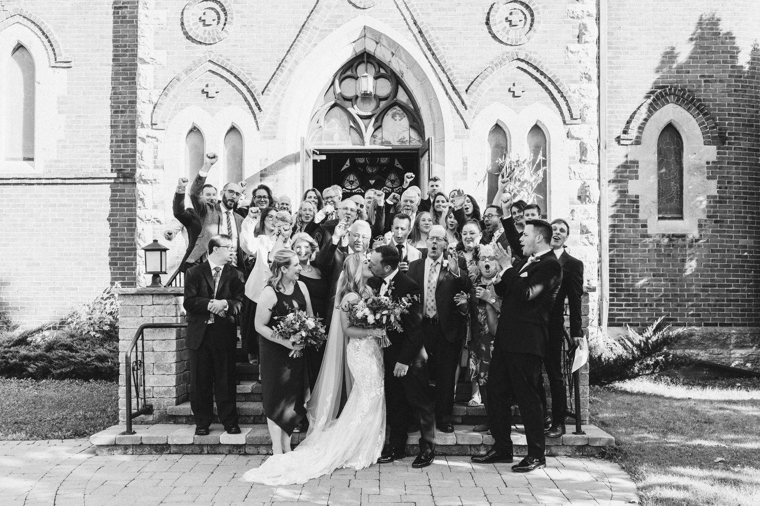 Large wedding party gathered on church steps for a celebratory group photo.