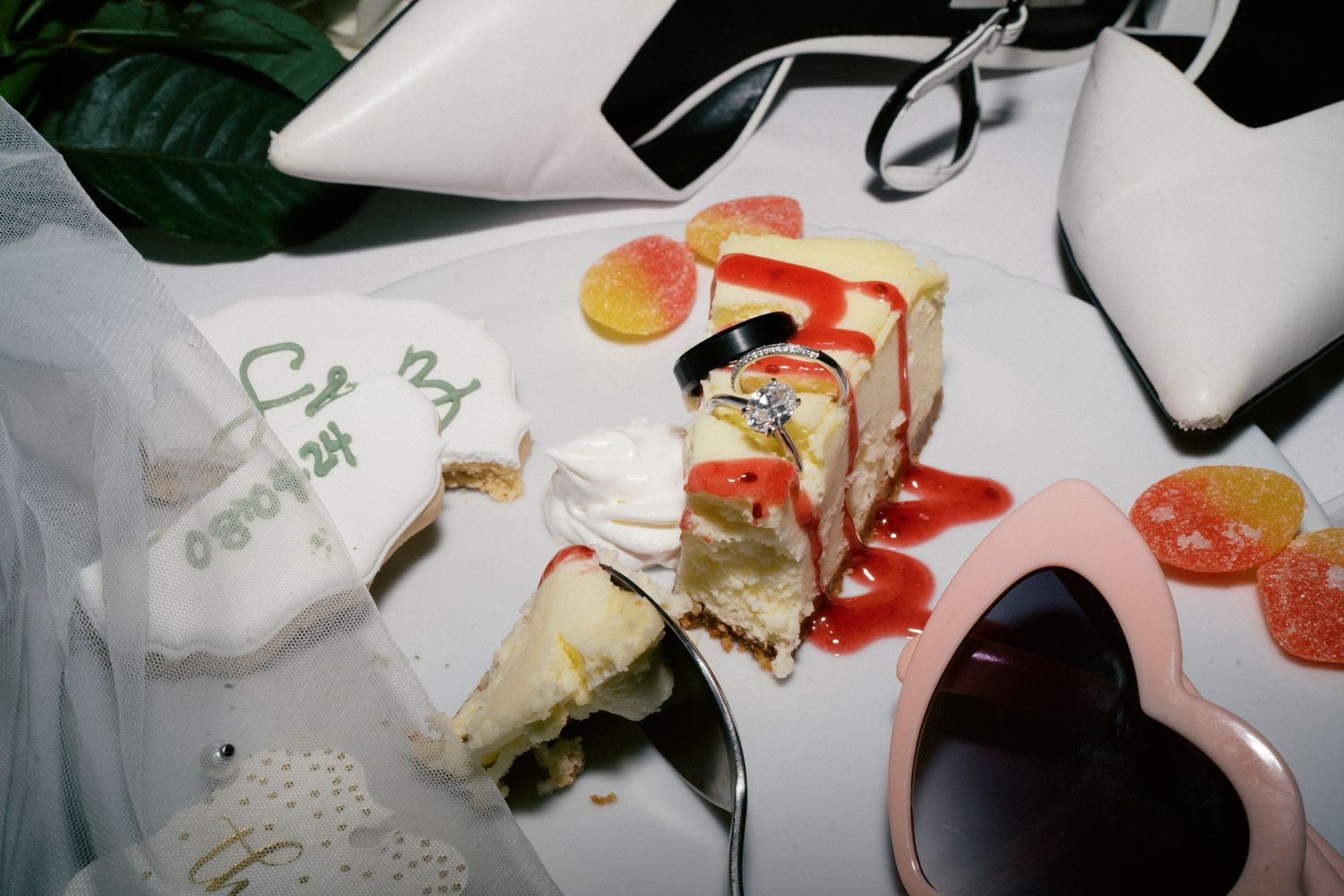 Messy wedding dessert plate with cheesecake, strawberry sauce, candy, and heart-shaped sunglasses on a white table.