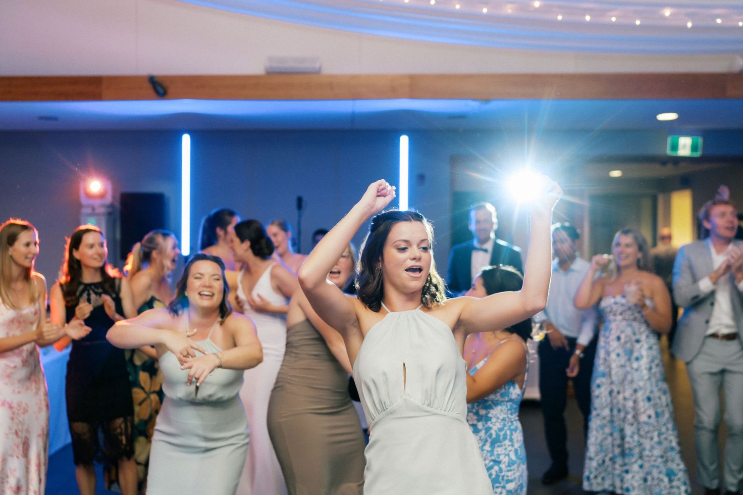 Bride dancing joyfully with arms raised at wedding reception, surrounded by guests on the dance floor.