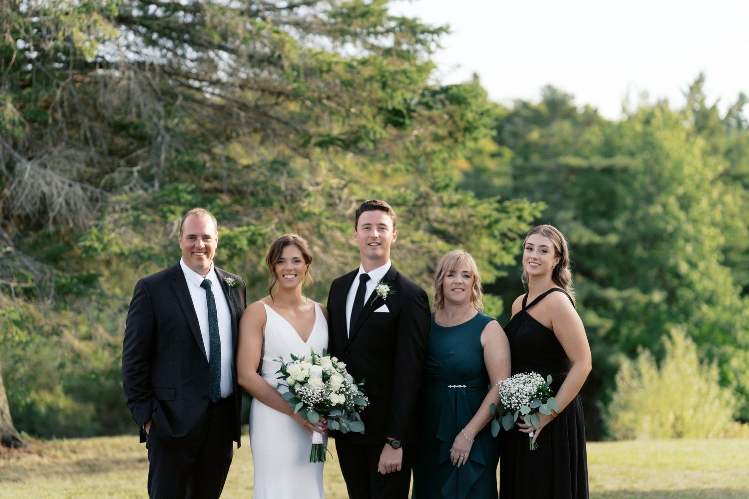 Wedding party in formal attire poses together outdoors against evergreen trees.