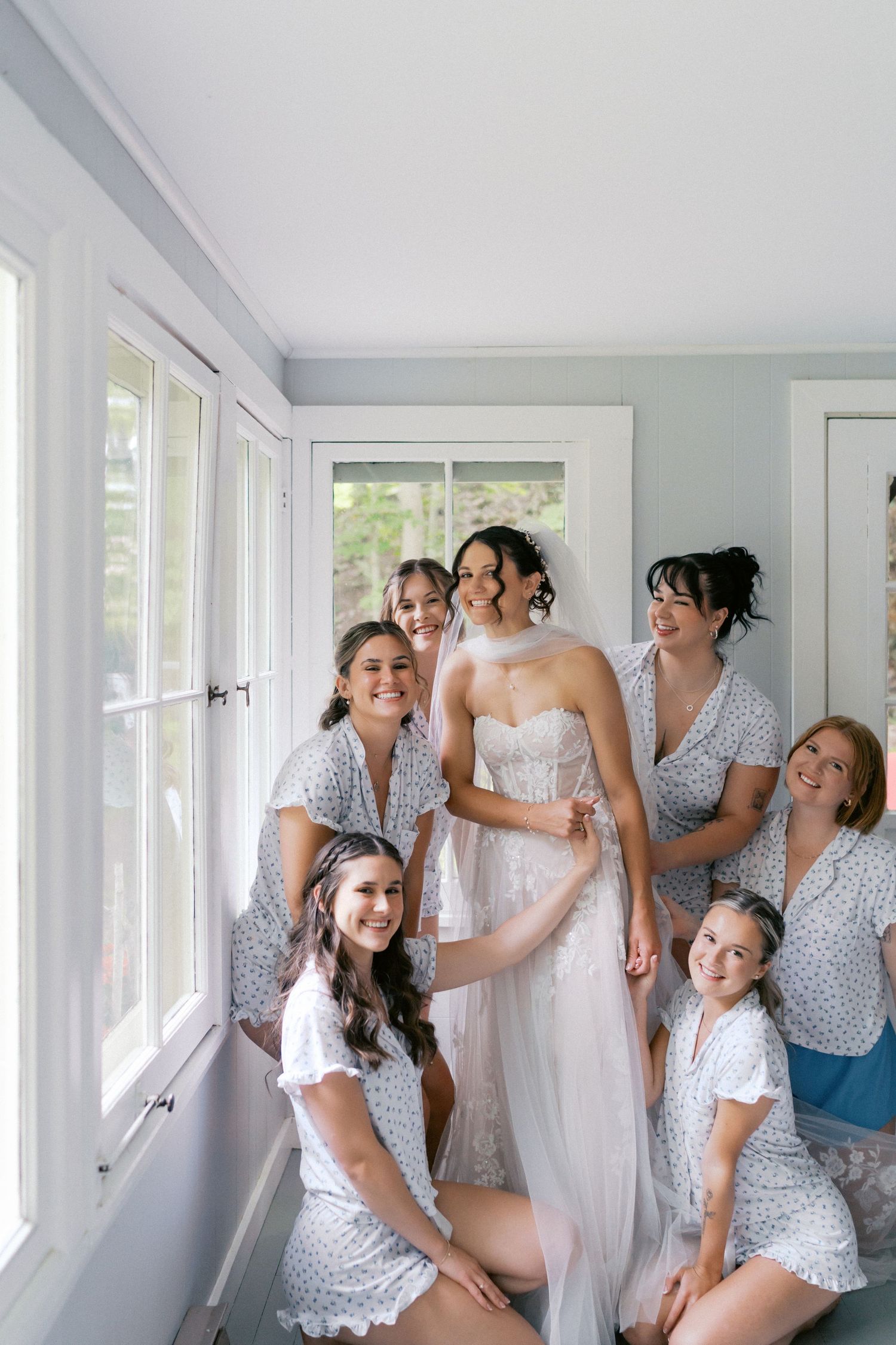 Bridesmaids in polka dot robes help with wedding preparations in a bright sunlit room.