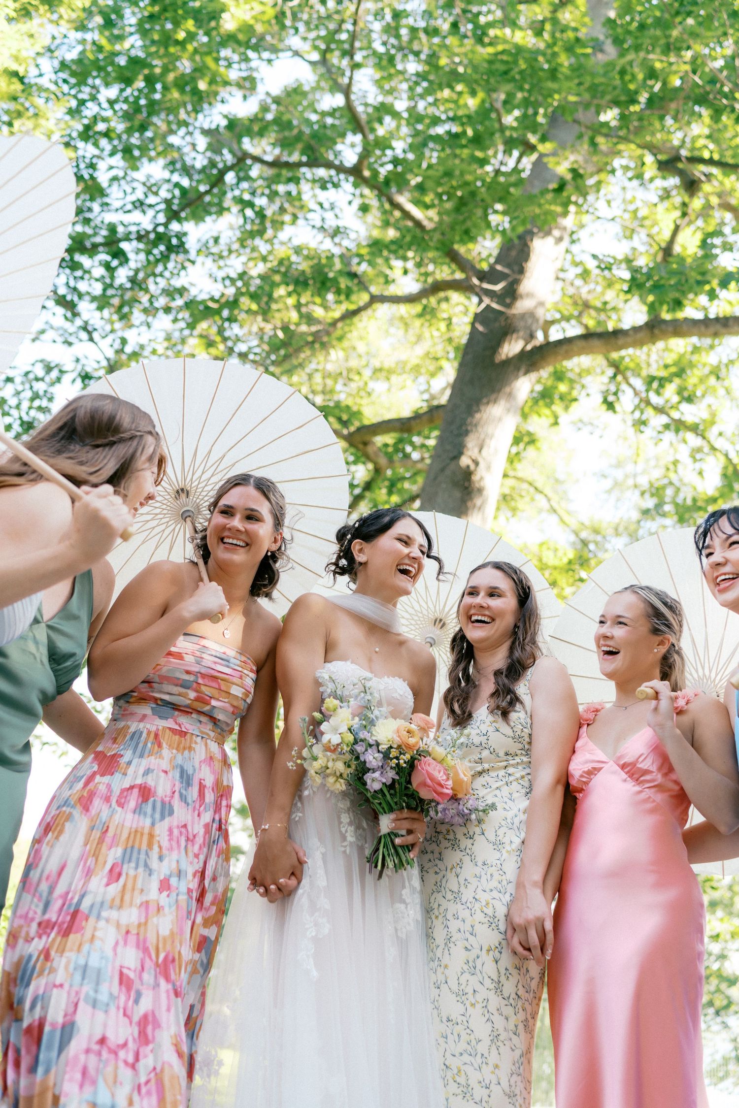 A joyful bridal party shares laughter under trees while holding white parasols.