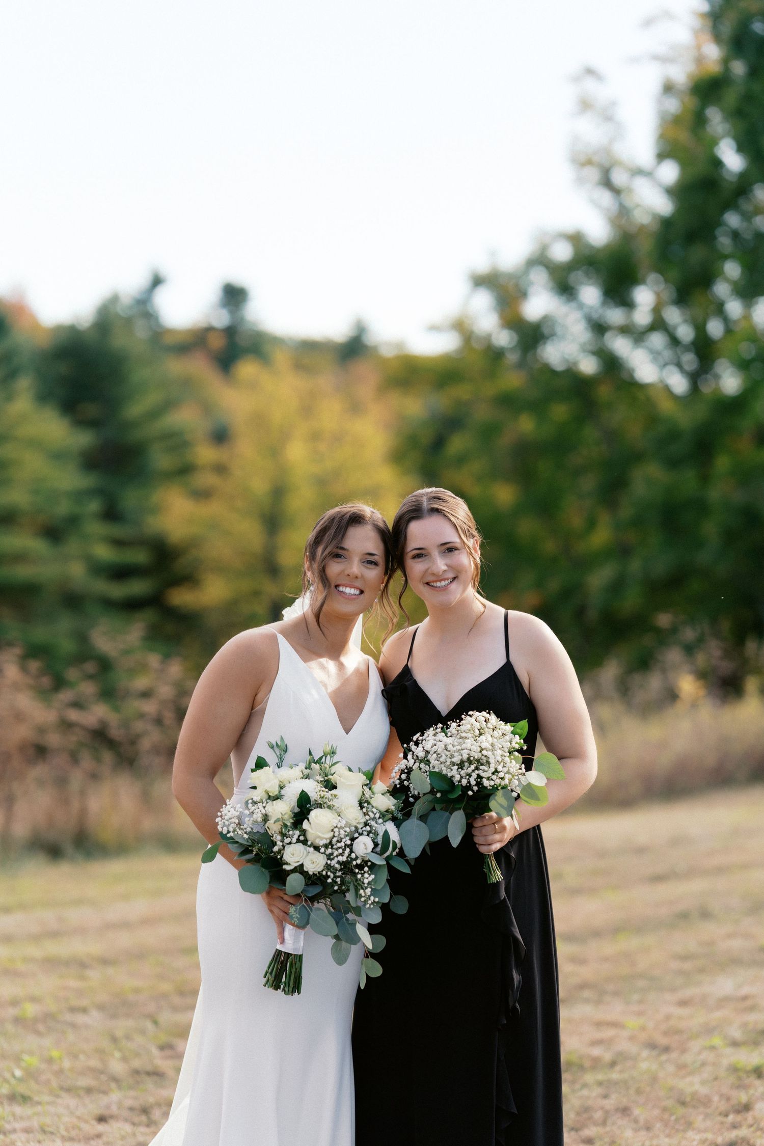 Two people in wedding attire share a joyful moment outdoors with white floral bouquets against an autumn backdrop.