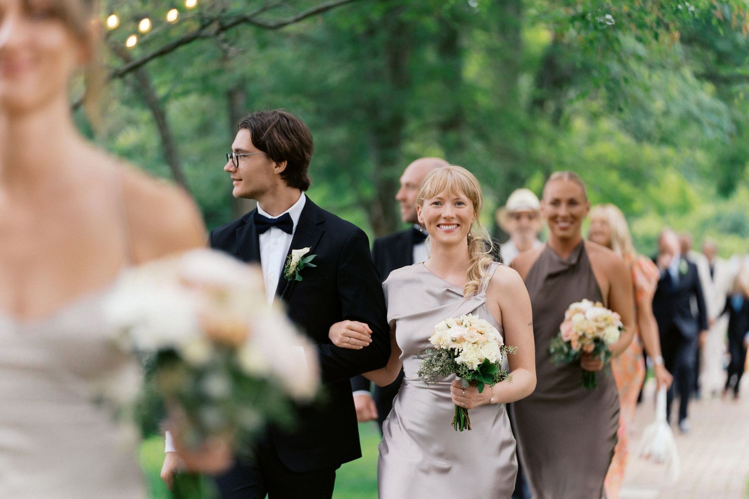 Wedding party members walk in a procession through a garden path carrying floral bouquets.