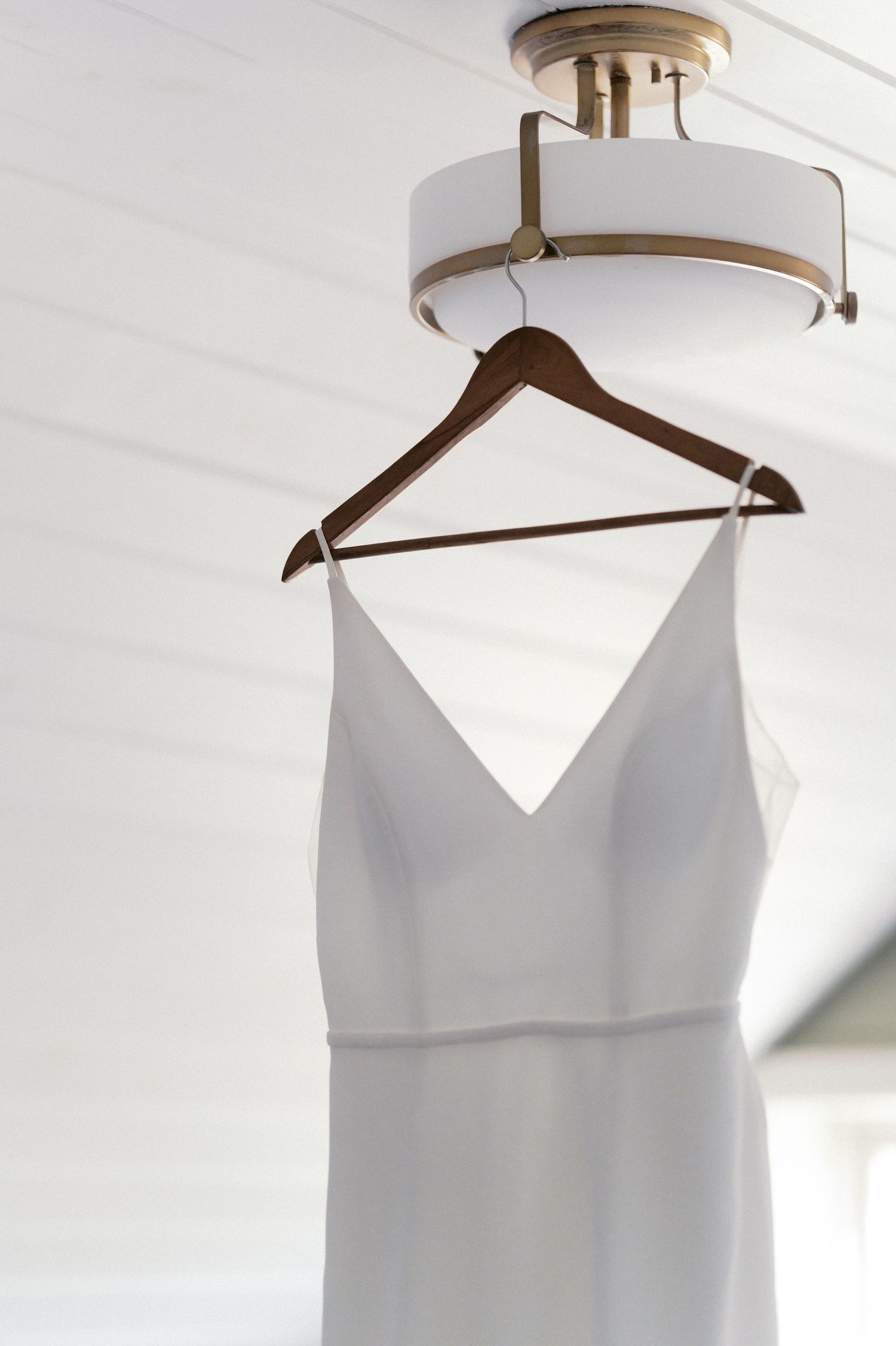 Elegant white wedding dress hanging on wooden hanger against light colored wall.