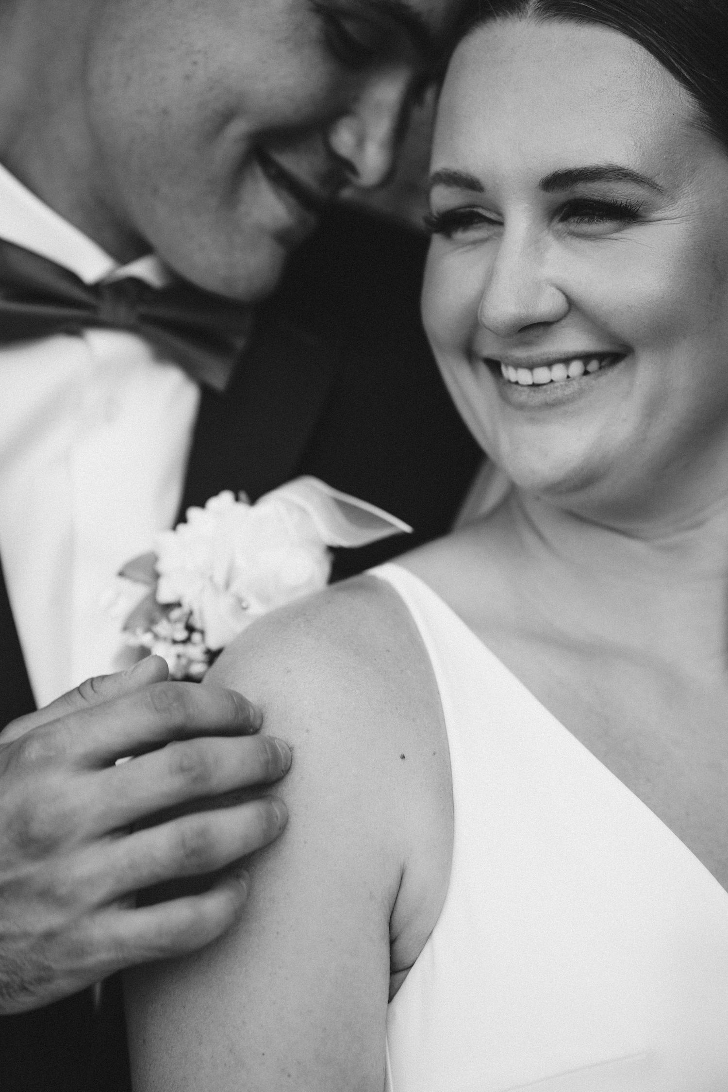 Black and white photo of a smiling bride and groom embracing, groom wearing a boutonniere on his lapel.