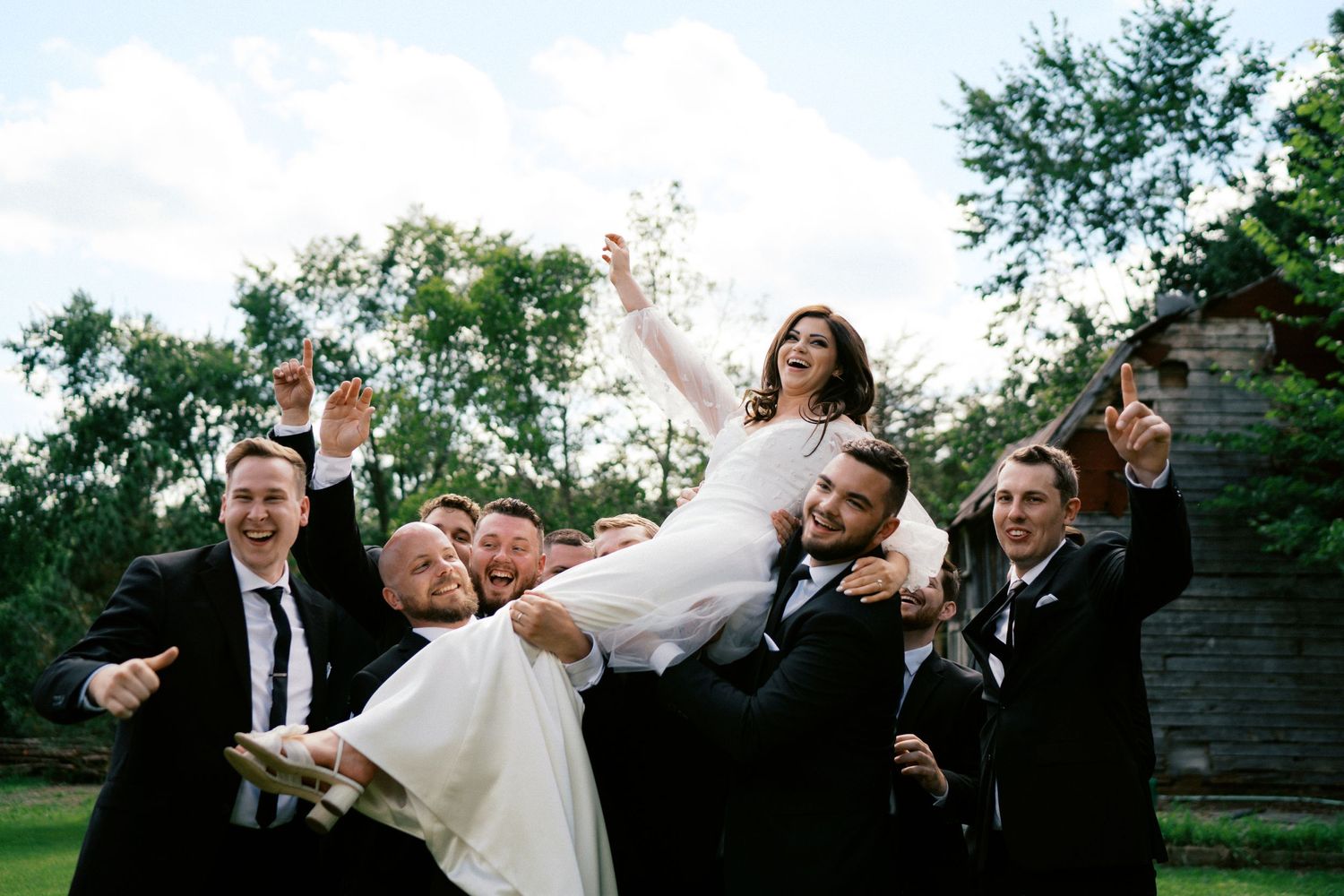 Wedding party lifts bride in celebratory pose on grassy lawn with joyful expressions.