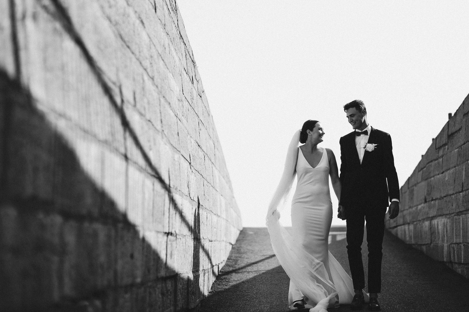 Elegant black and white photo of newlyweds walking along a concrete wall.