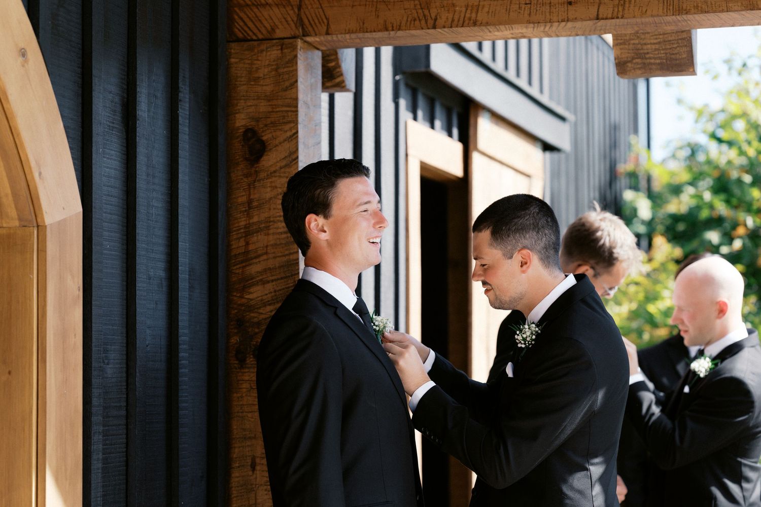 Wedding party in formal attire gathered by large black barn-style building.