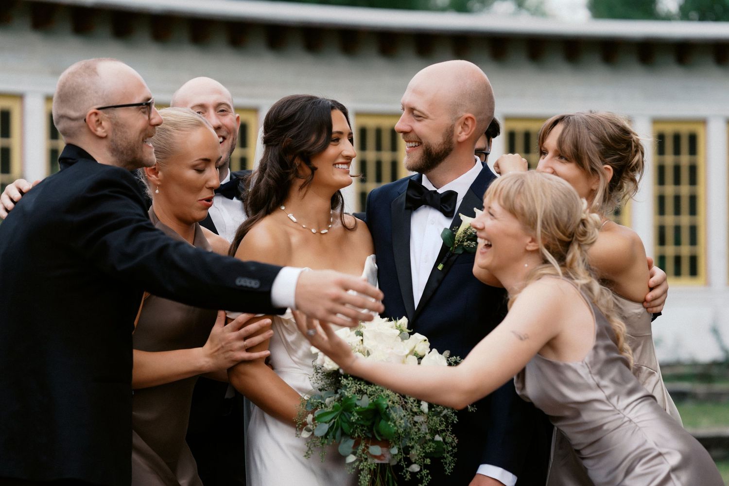 Wedding party members embrace and celebrate during formal photos outdoors.