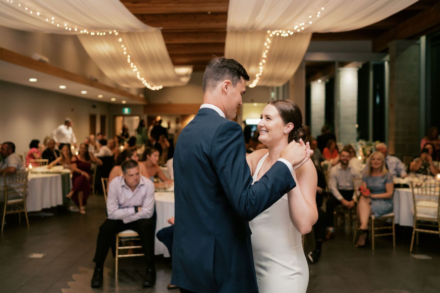 Bride and groom share their first dance at elegant wedding reception with fairy lights and guests seated behind.