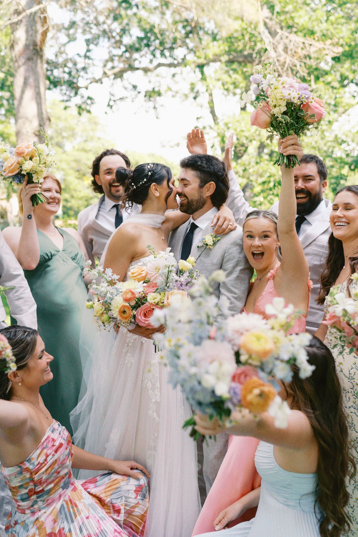 Wedding party celebrates joyfully outdoors with colorful bouquets and festive expressions.