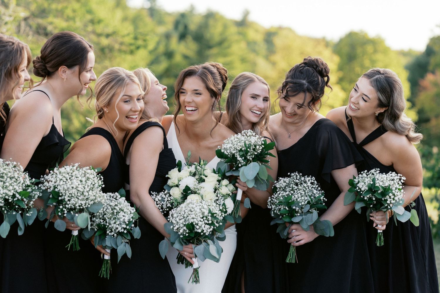 Bridesmaids in black dresses hold white flower bouquets while standing in a row outdoors.