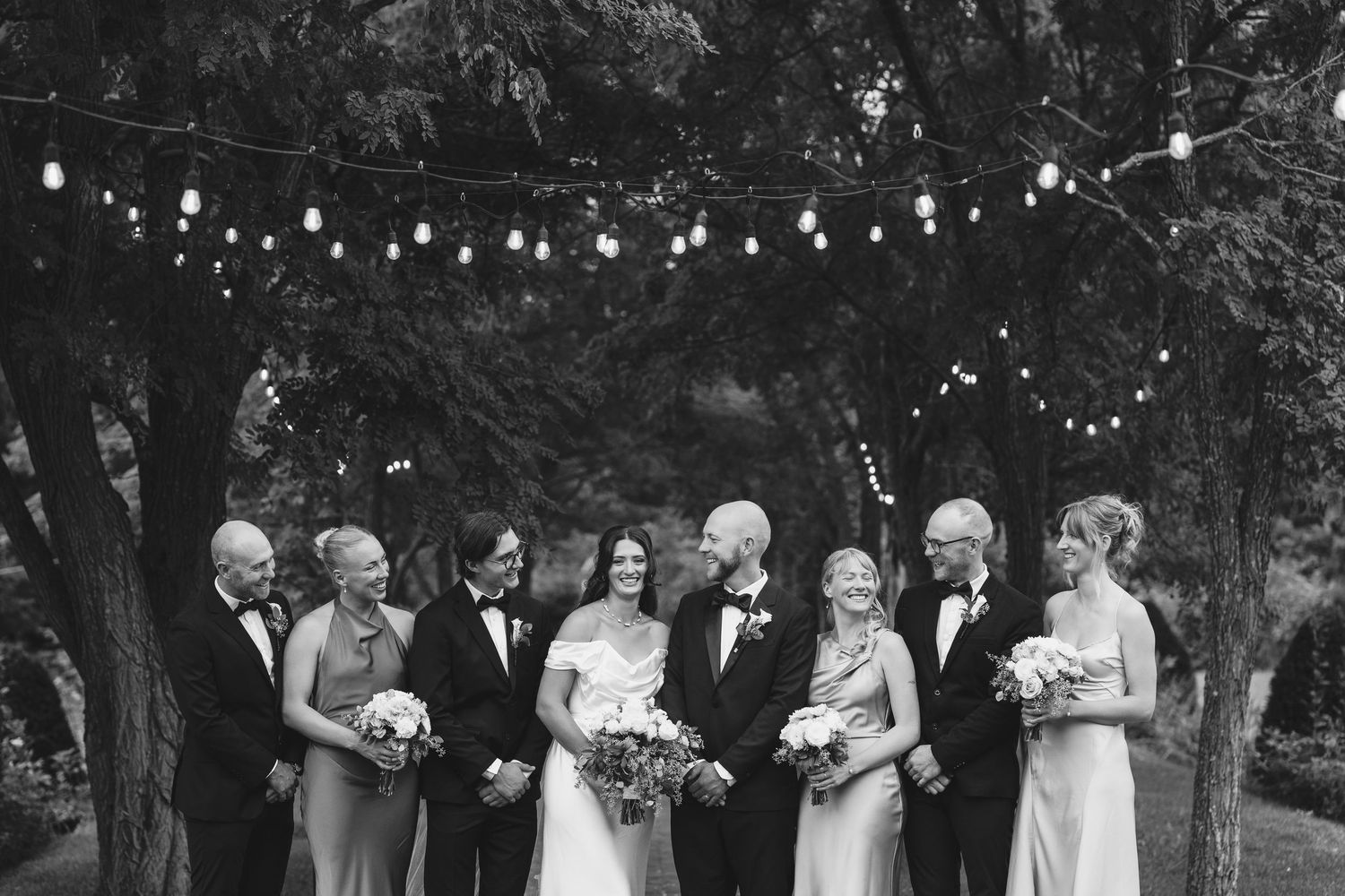 Black and white photo of a wedding party gathered outdoors under string lights.