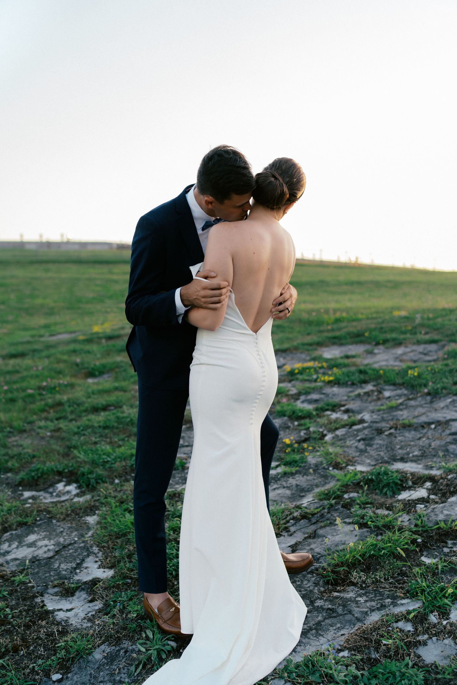 A bride and groom share a romantic embrace in an open field, with the bride wearing a backless white gown.