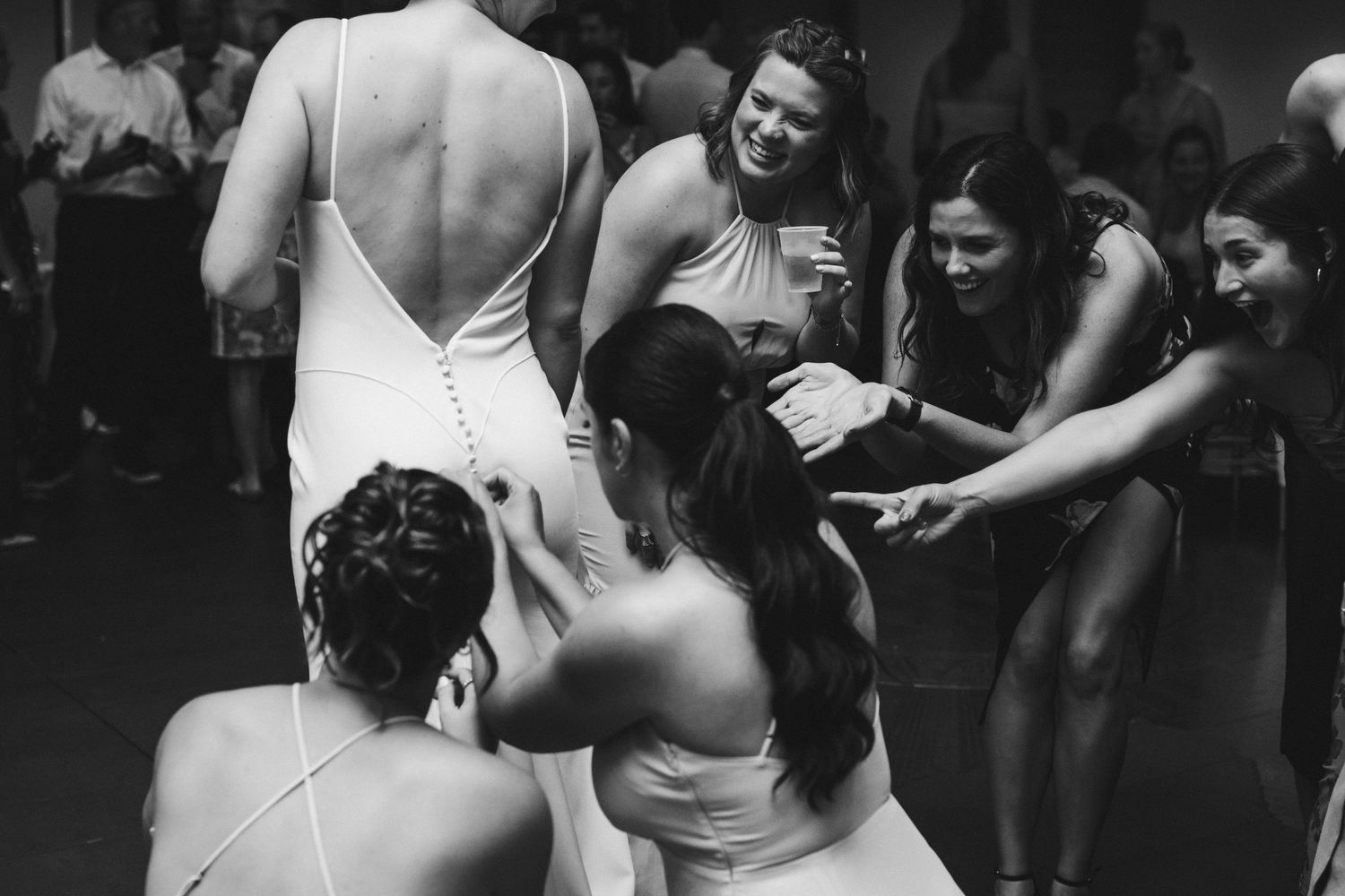 Bridesmaids hugging bride in white backless gown on dance floor at wedding reception, black and white photo.