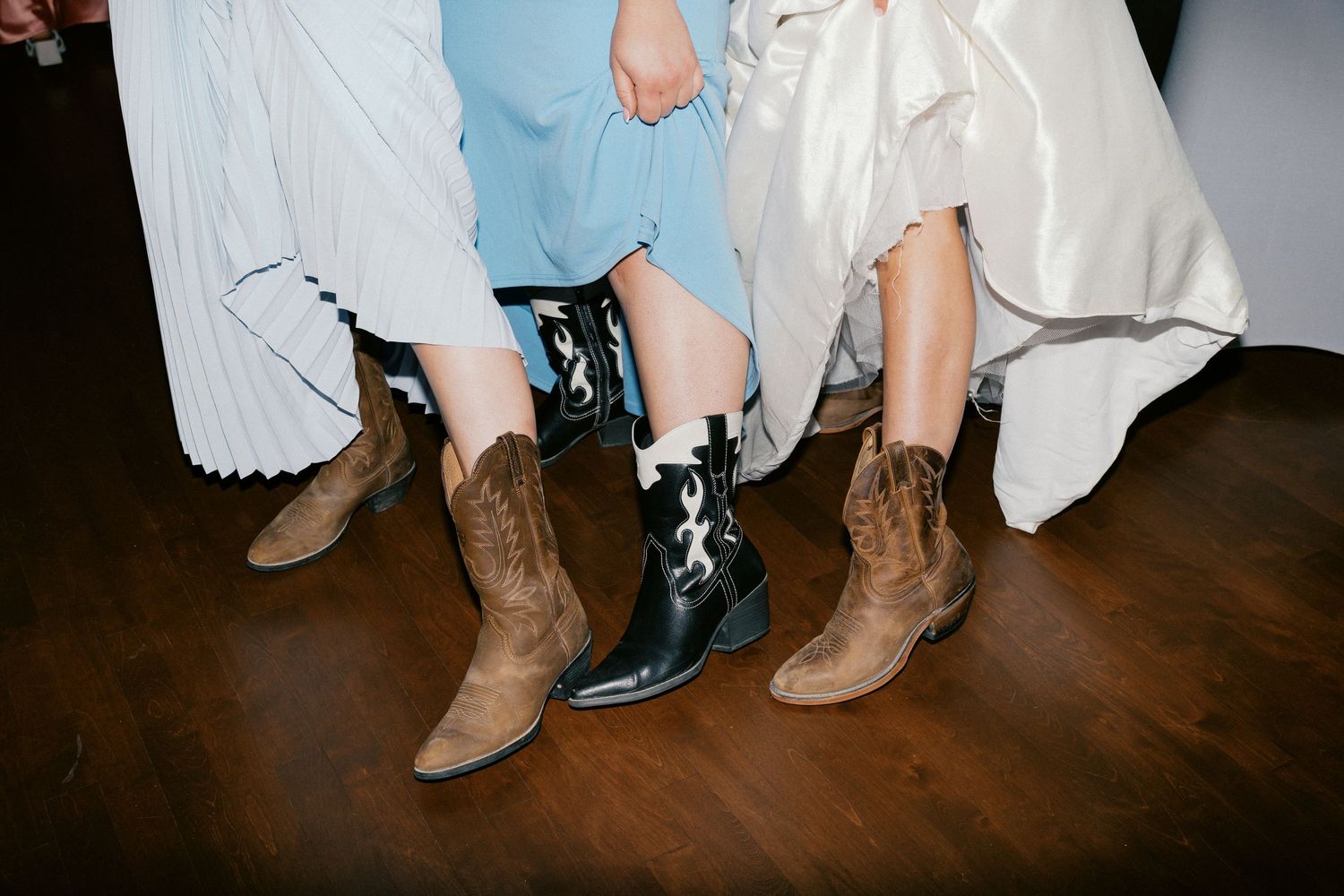 A group of cowboy boots worn by wedding party members lined up on a dark wooden floor.