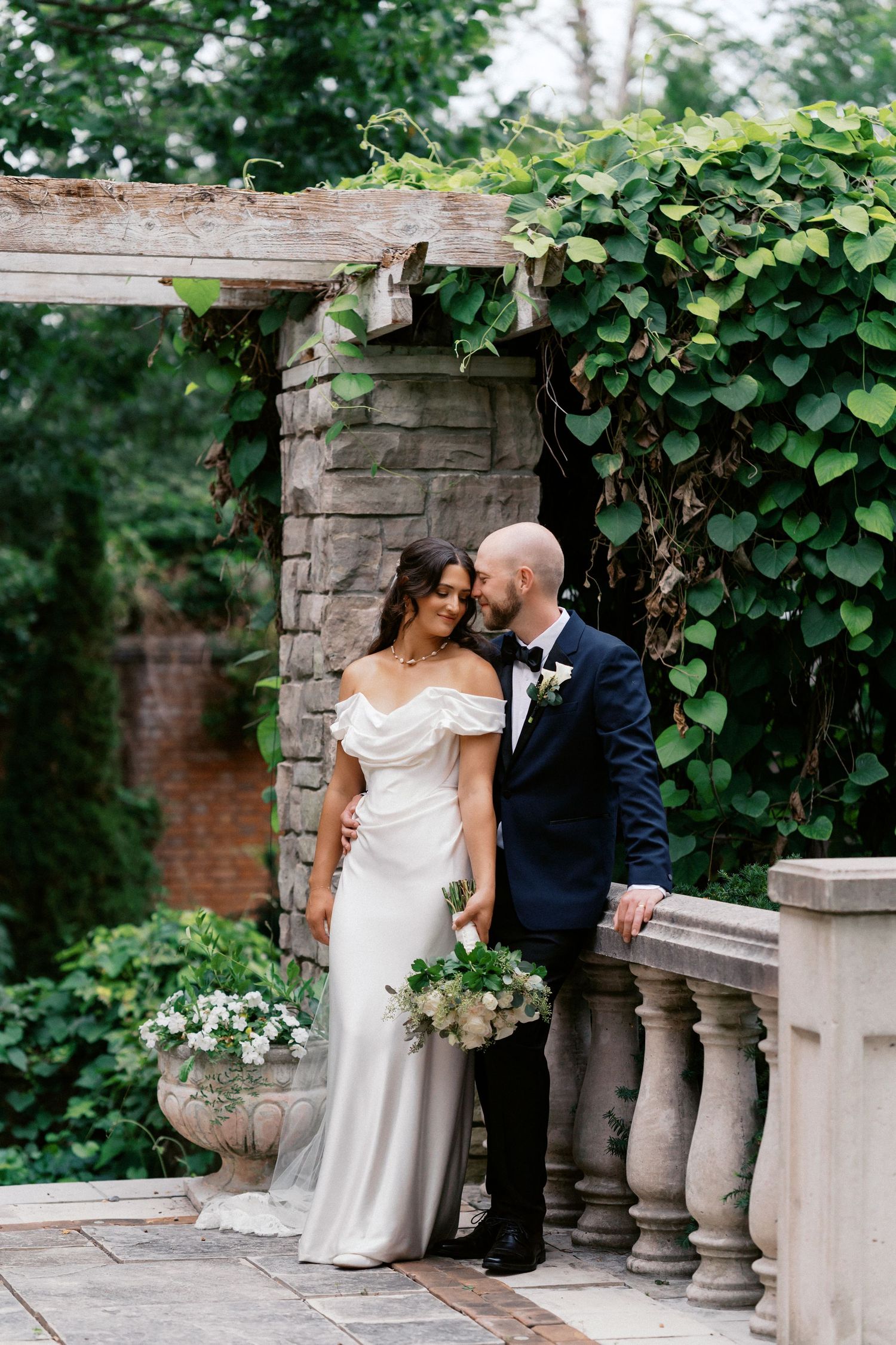 Romantic wedding couple poses along stone balustrade covered in climbing vines.