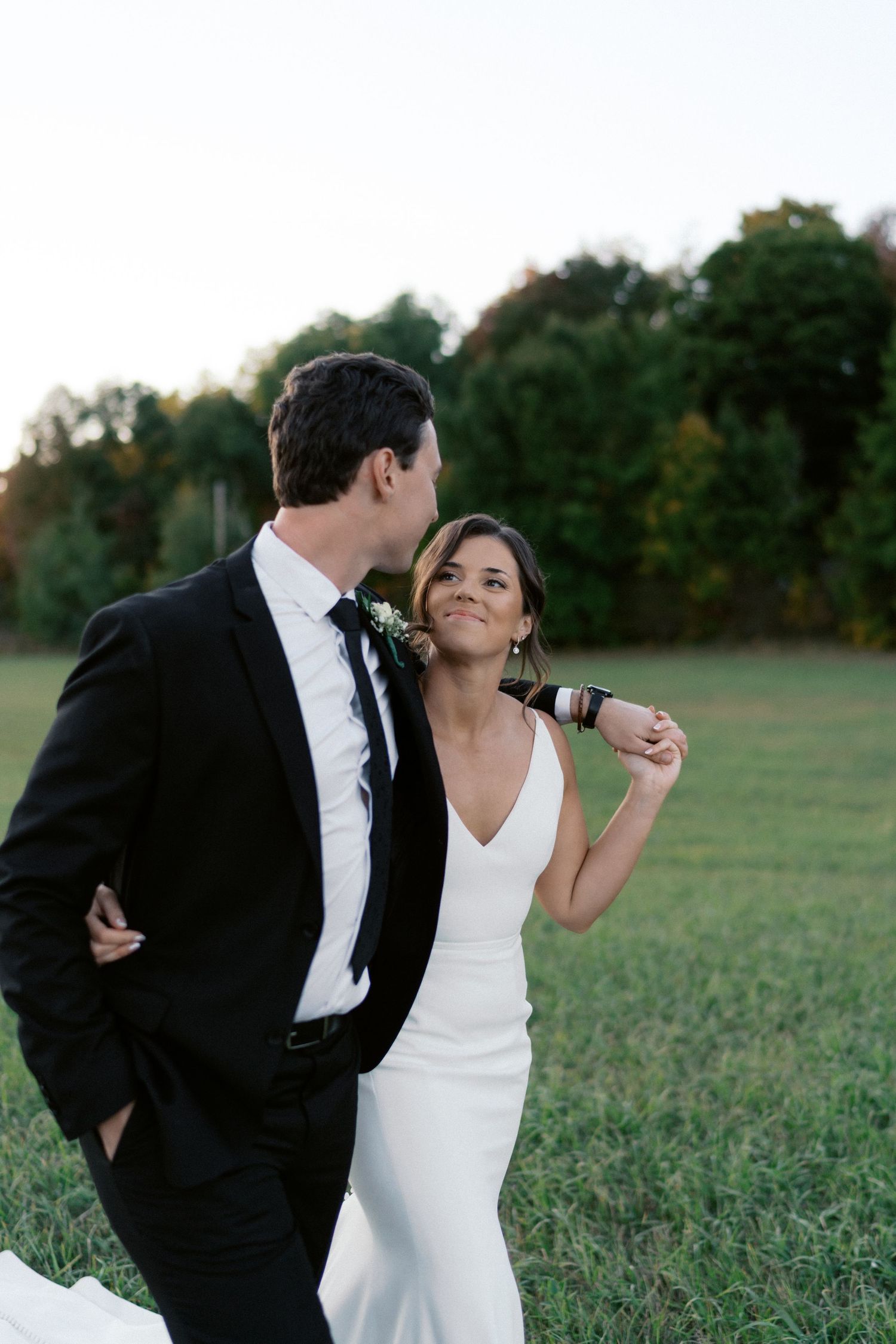 A couple in wedding attire playfully poses together in a green field at sunset.